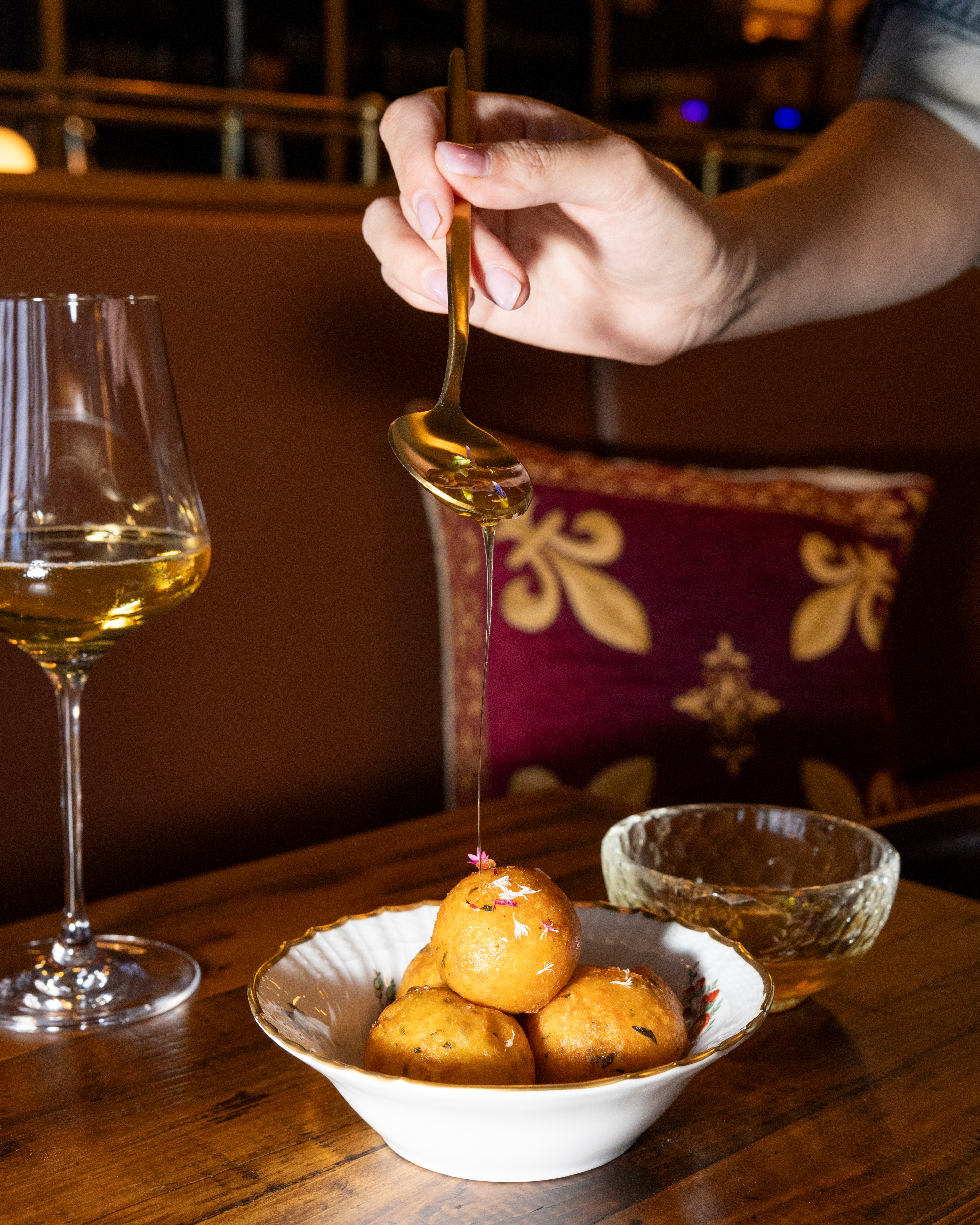 A hand drizzles honey over a bowl of fried dough balls; a wine glass and another dish are nearby on the wooden table, with a decorative pillow in the background.