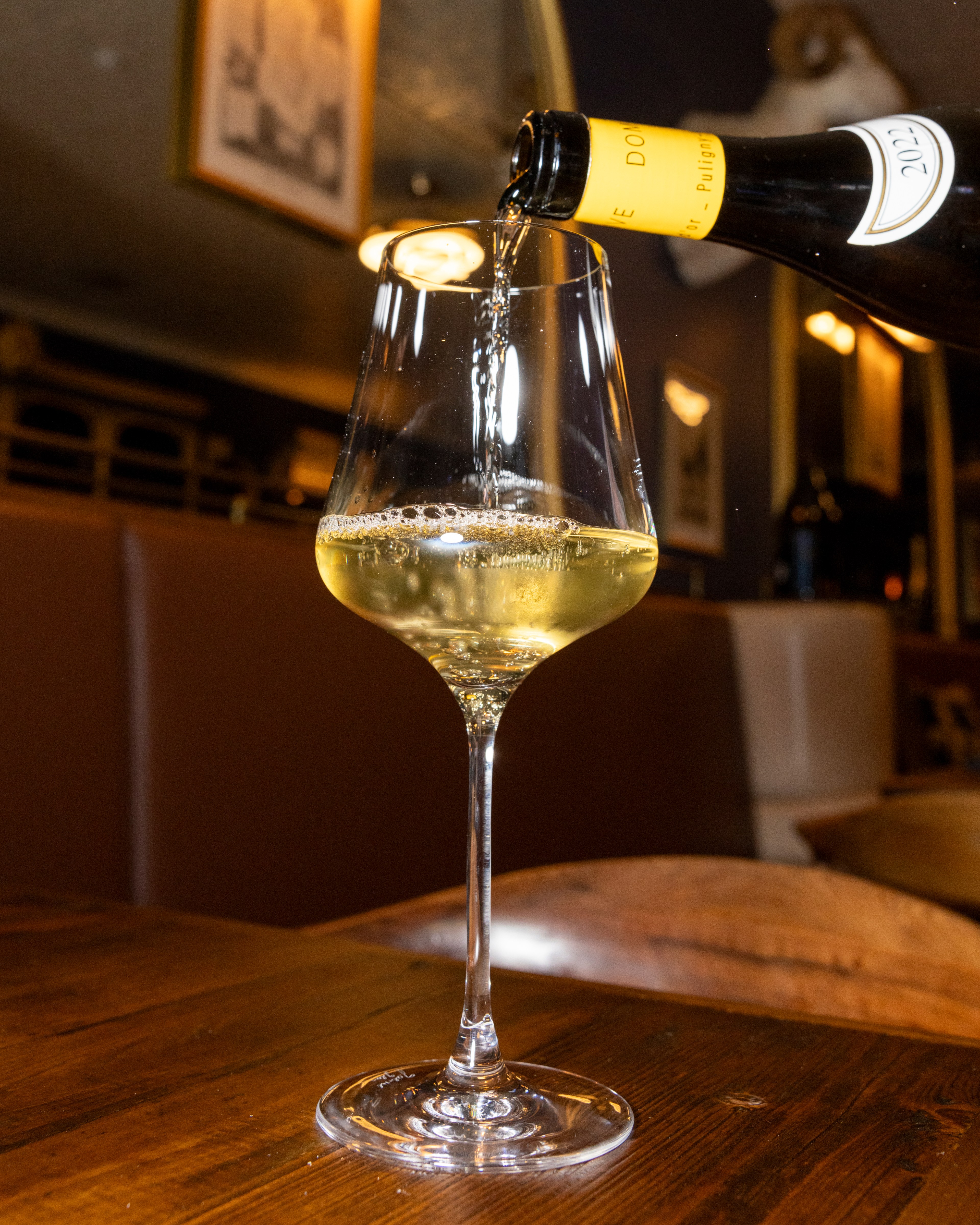 A wine bottle is pouring a light-colored wine into a clear, elegant wine glass on a wooden table in a dimly-lit, cozy setting.