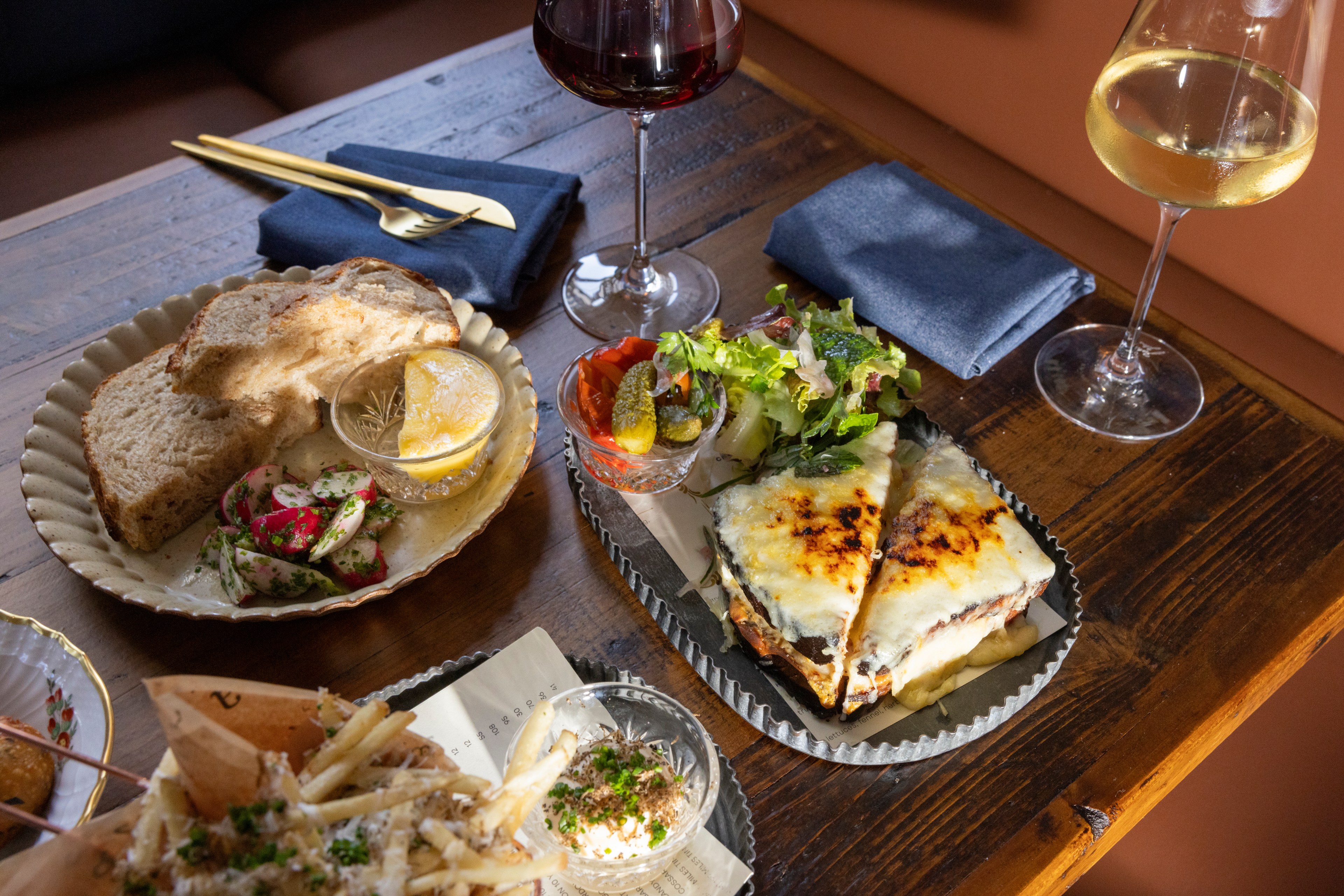 This image shows a wooden table with a meal including bread, salad, seasoned fries, a cheesy sandwich, and two glasses of wine—one red, one white.