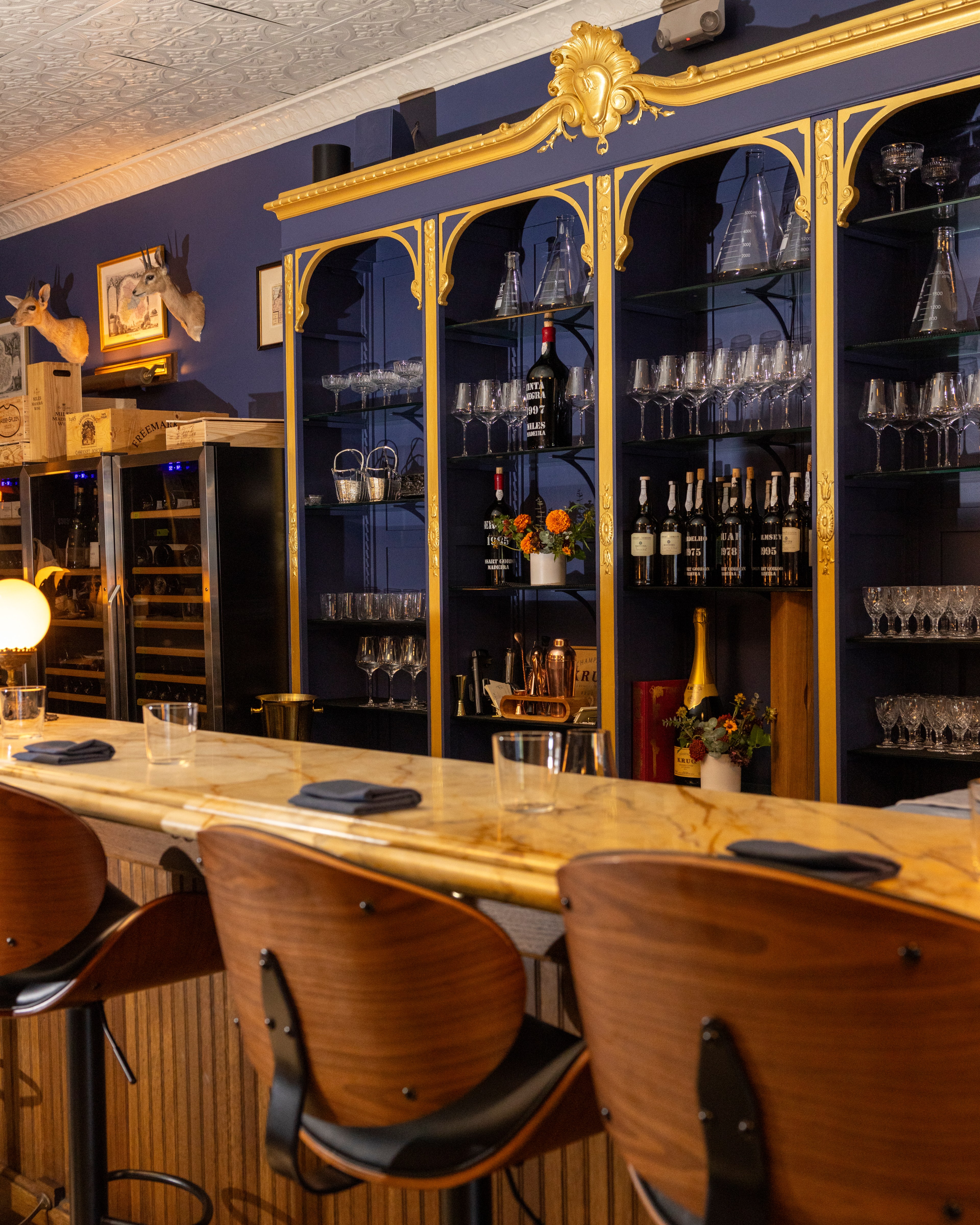 This image shows a stylish bar with wooden chairs, a marble countertop, and neatly arranged glassware on shelves. The backdrop includes ornate, gold-accented cabinets.