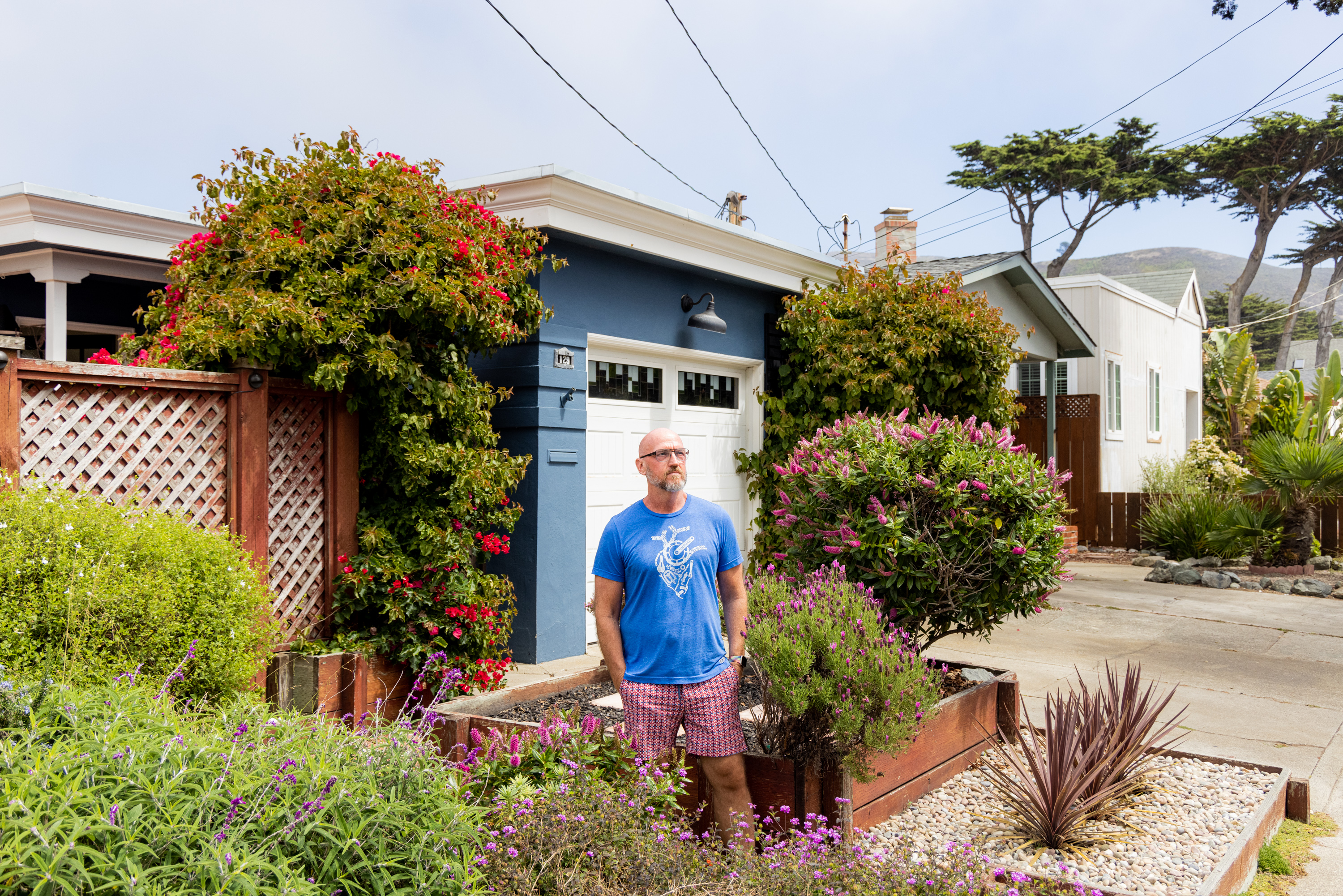 A man in a blue shirt and pink shorts stands in front of a blue house with a white garage door, surrounded by lush green and flowering plants on a sunny day.