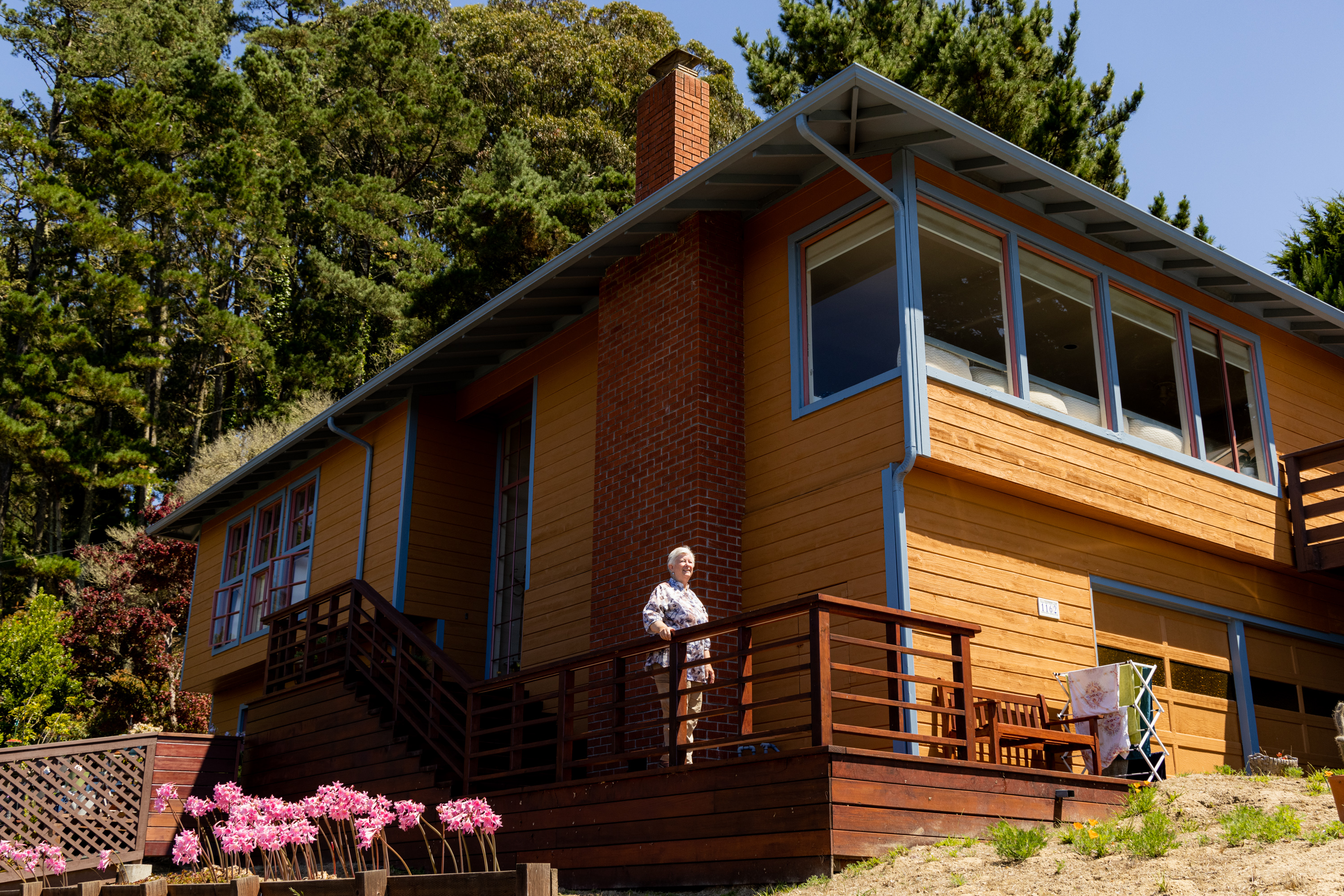 An older woman stands on a wooden deck attached to a large, yellow and blue two-story house with big windows. Pink flowers bloom along the fence, and a green forest is in the background.