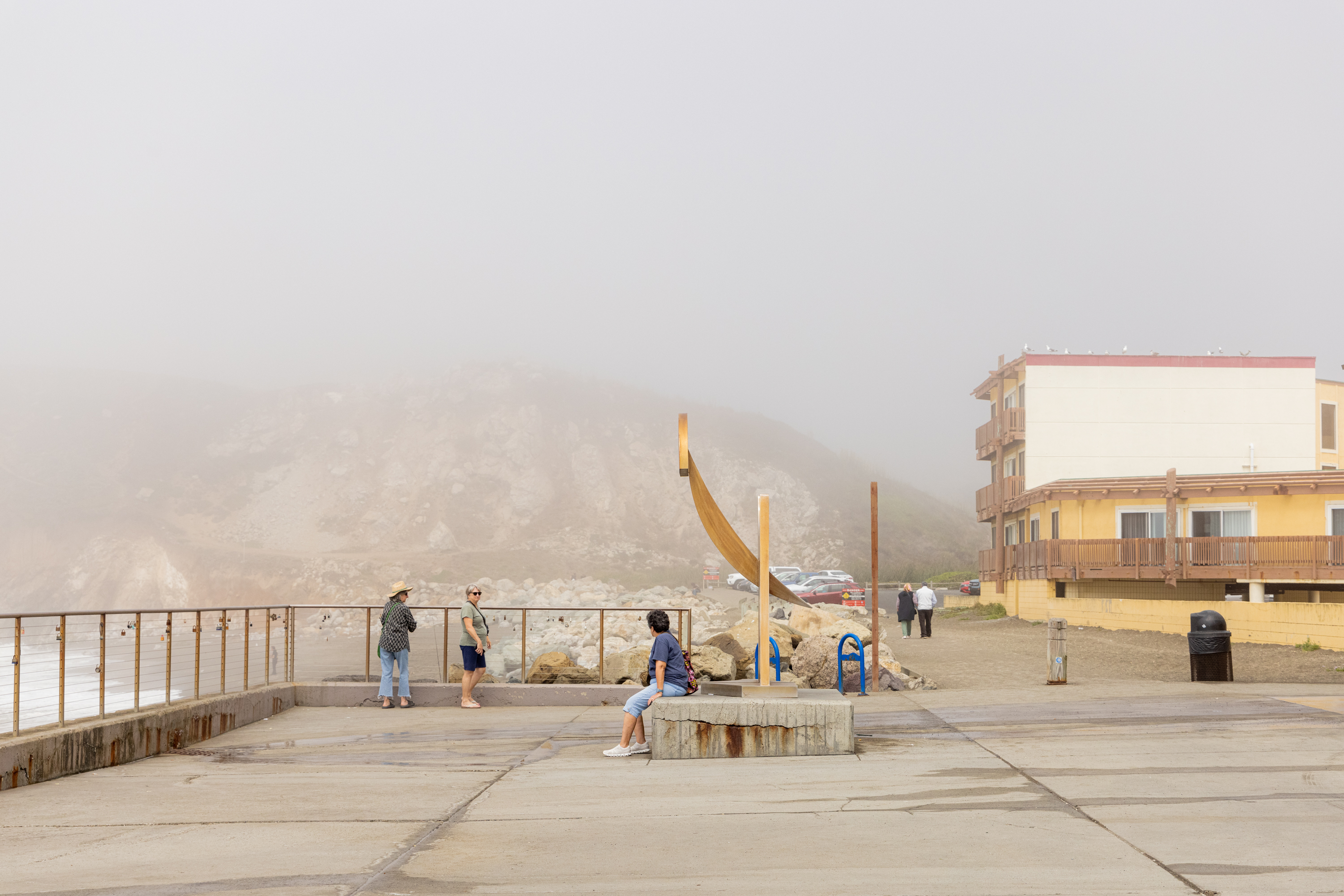 People are walking and sitting near a beachfront area with a foggy background, featuring a rocky hillside and a yellow multi-story building.