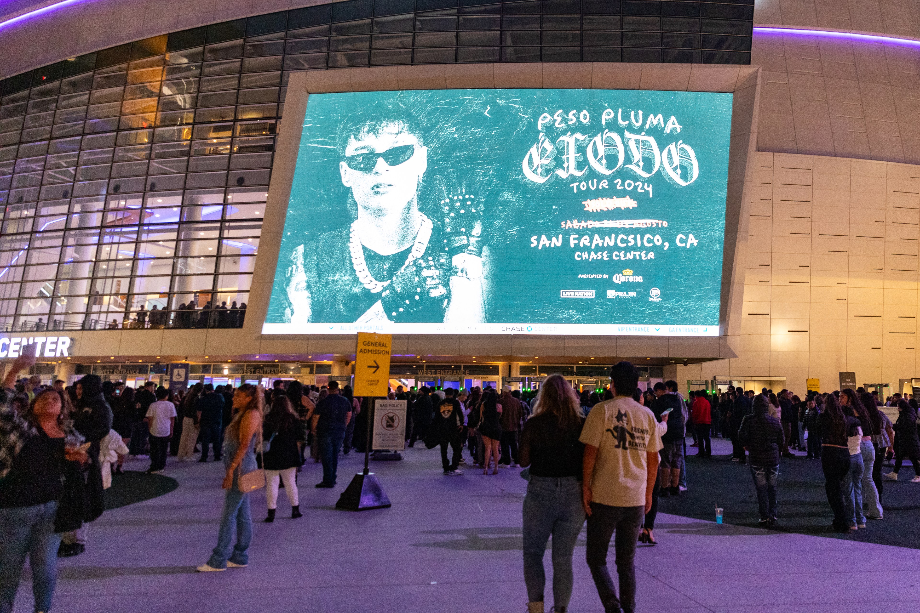 A crowd gathers outside a venue at night, with a large screen above showing an image of a person in sunglasses and chains, advertising the &quot;Peso Pluma Tour 2024&quot; in San Francisco.