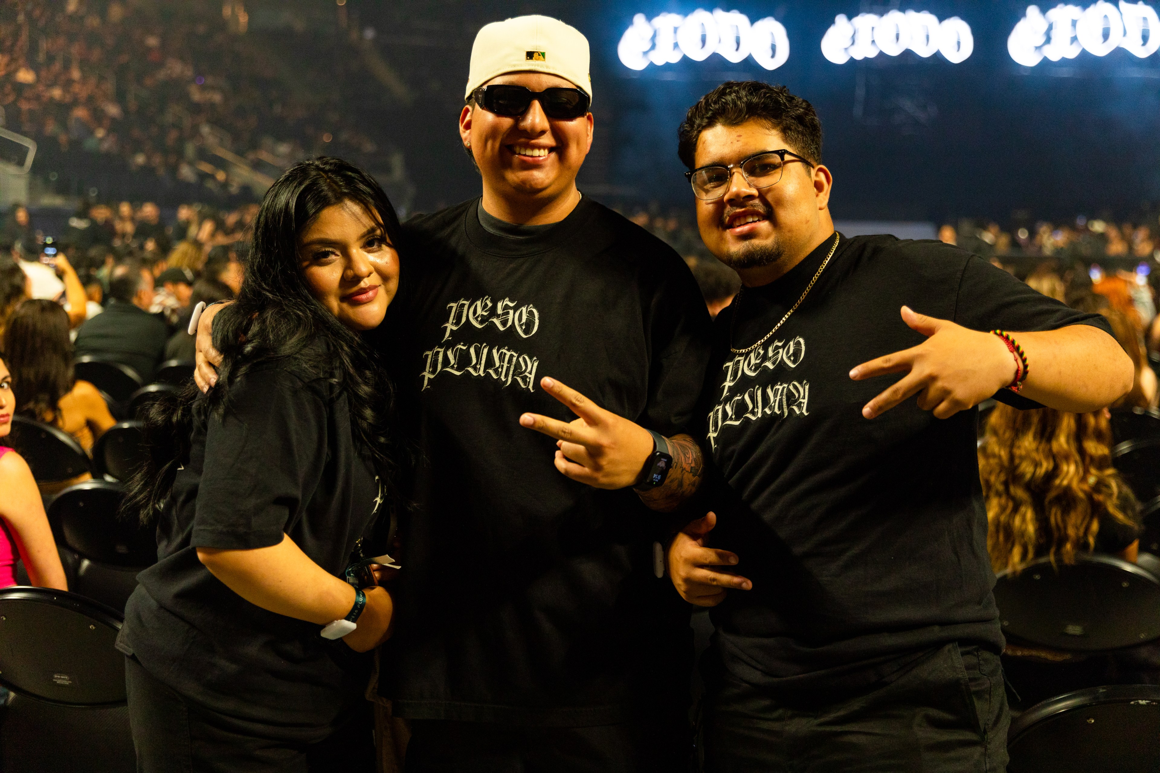 Three smiling individuals in black &quot;Peso Pluma&quot; shirts pose in an indoor concert venue with a filled audience. The background features a lit sign.