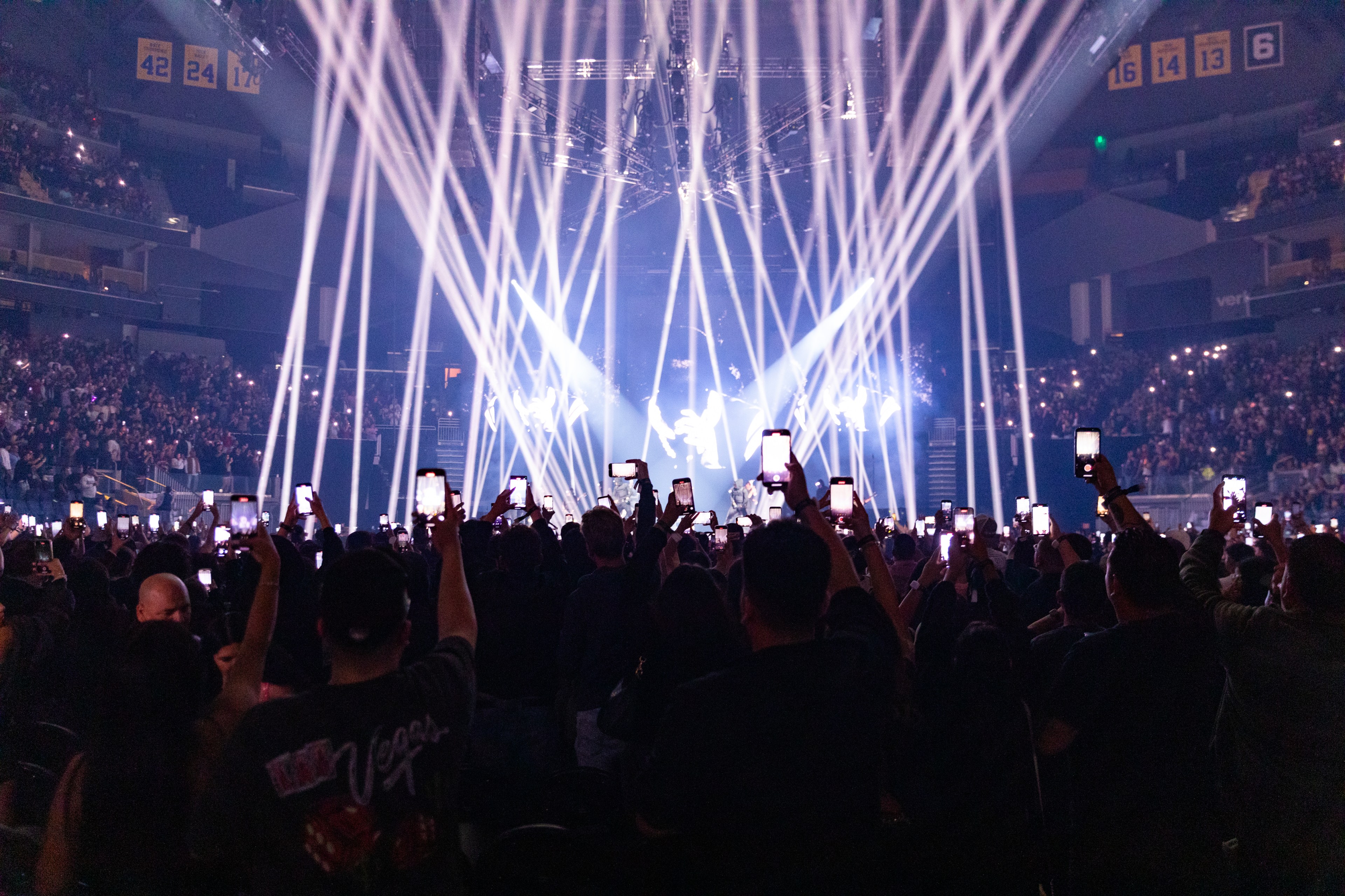 A large concert with bright stage lights beaming into the audience. Many attendees are holding up smartphones to record or photograph the performance.