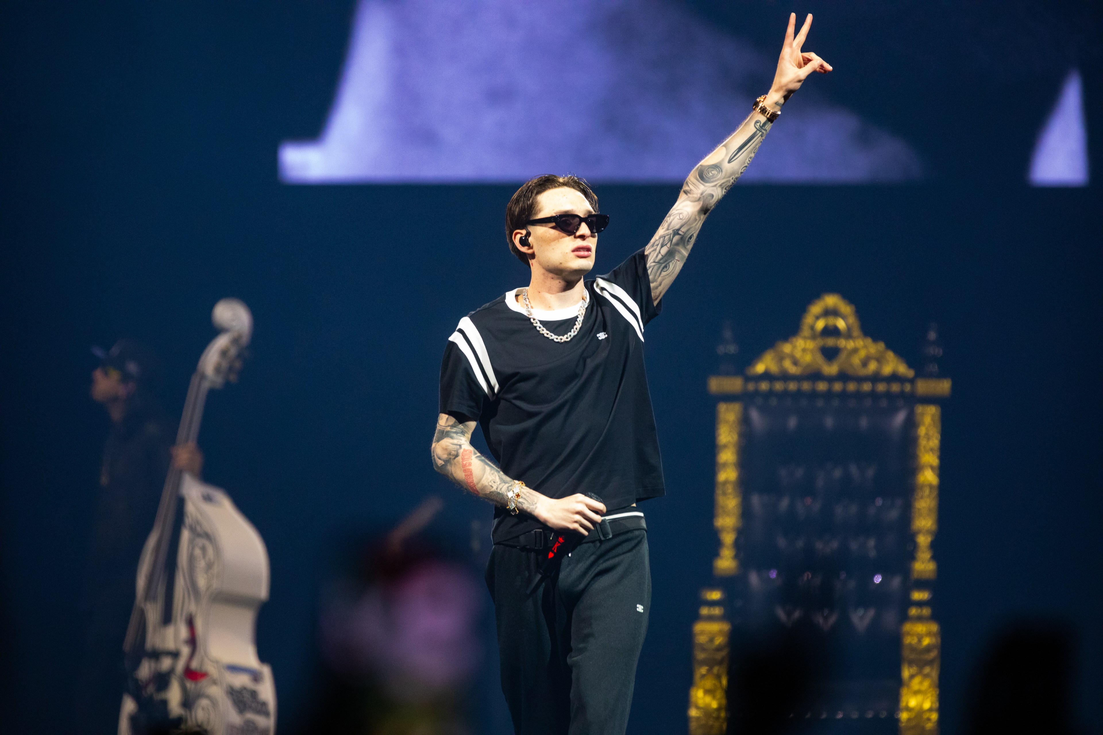 A tattooed man wearing sunglasses, a black shirt, and black pants performs on stage, raising his hand with a peace sign. A musician with a double bass and a throne are in the background.