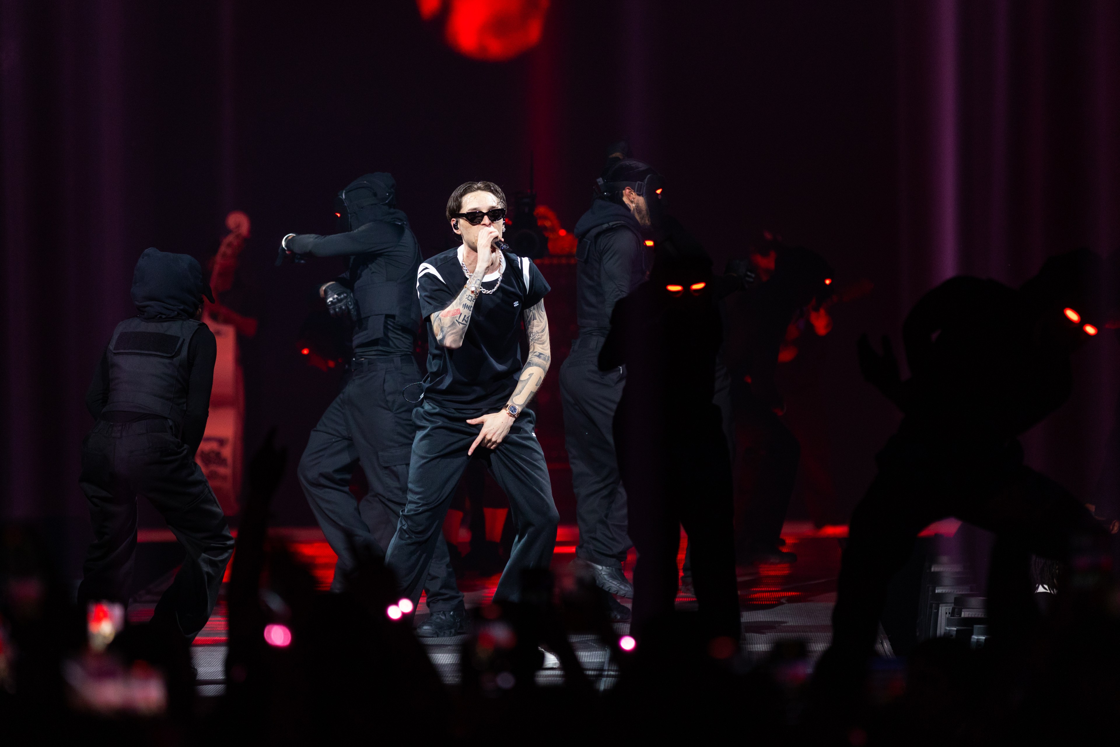 A performer in sunglasses and black attire sings into a microphone while surrounded by masked and hooded dancers under red stage lights.