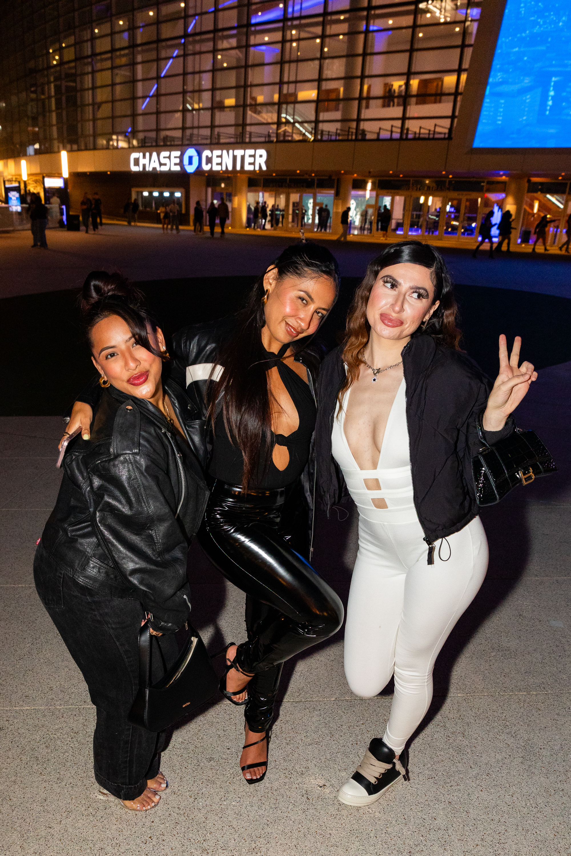 Three women in stylish outfits pose happily in front of the illuminated Chase Center at night, with one flashing a peace sign.