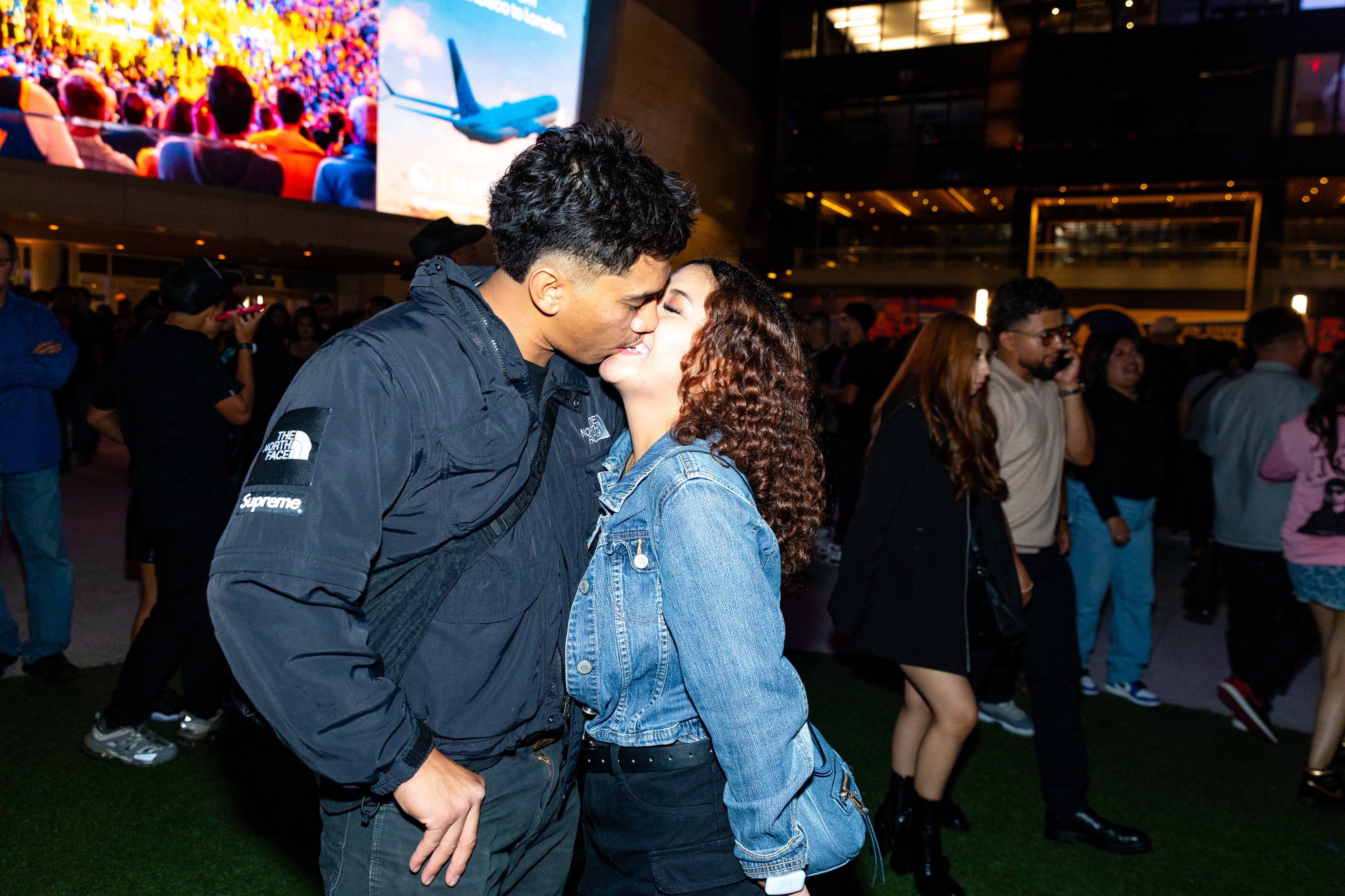 A couple kisses passionately in an outdoor crowd at night; the man wears a black jacket, the woman a blue denim jacket. Bright screen and people in the background.