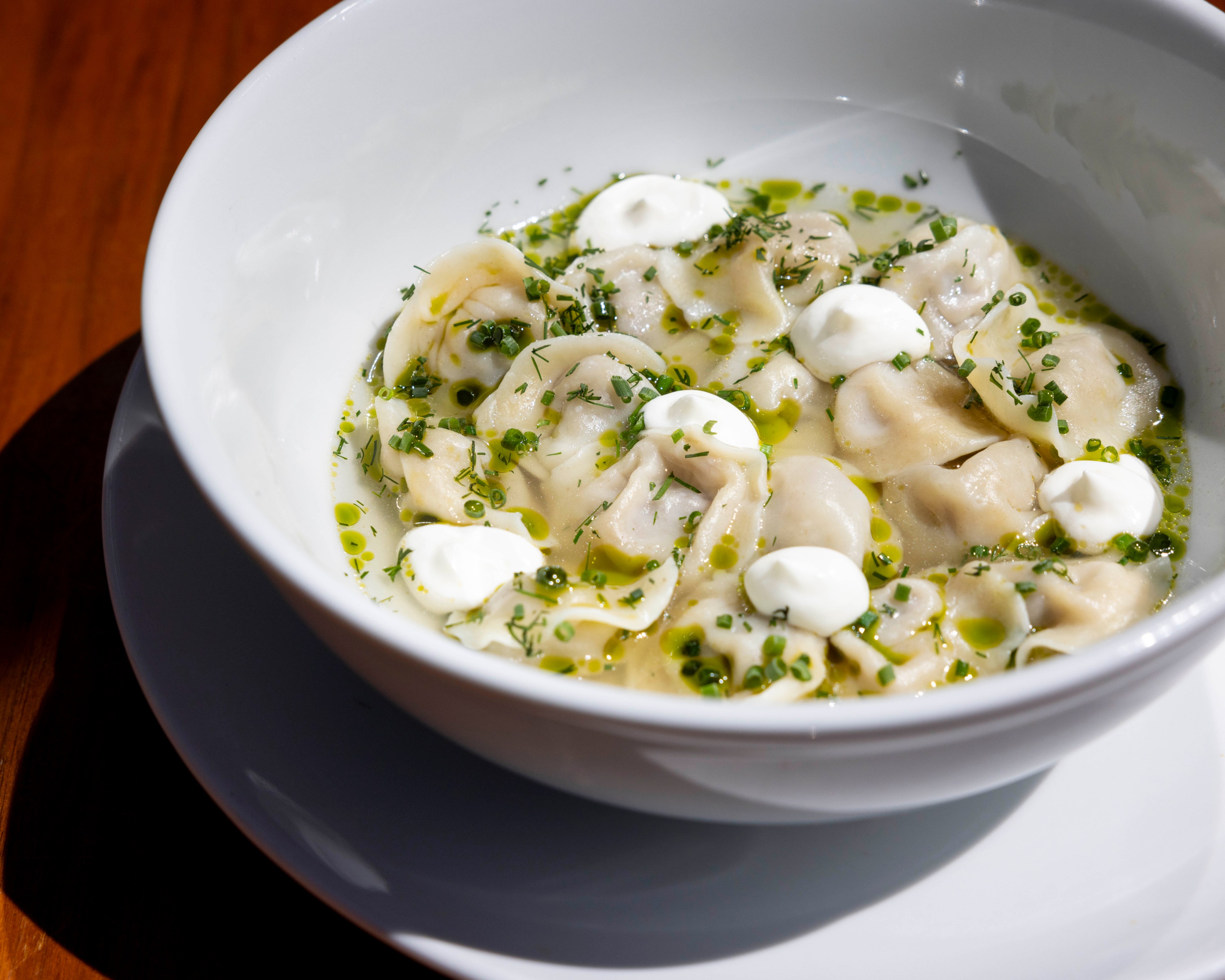 A white bowl filled with dumplings in a light broth, topped with small dollops of sour cream and sprinkled with fresh herbs.