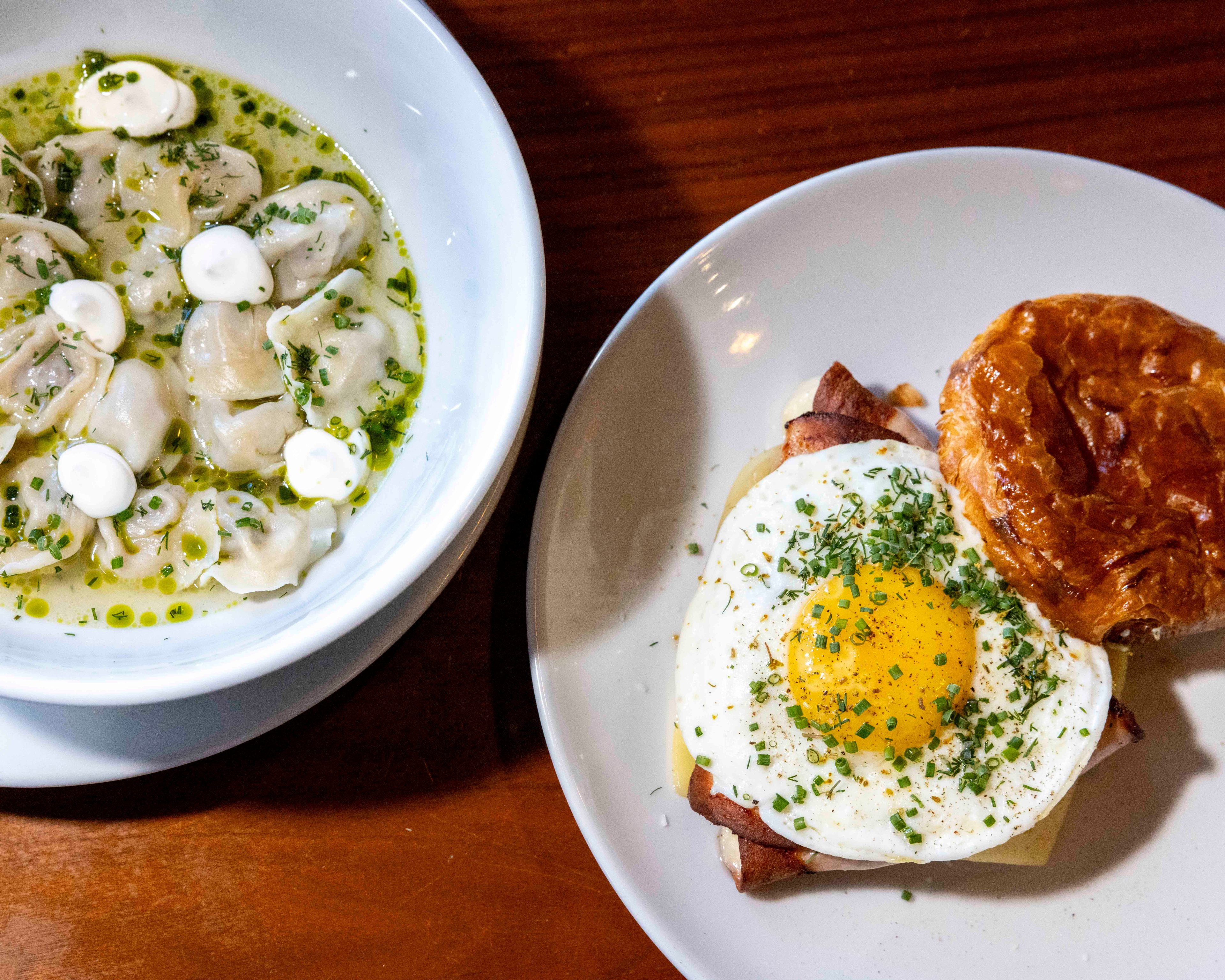 The image shows a bowl of dumplings in a light green broth with white dollops and herbs, and a plate with a sunny-side-up egg on toast and a pastry.