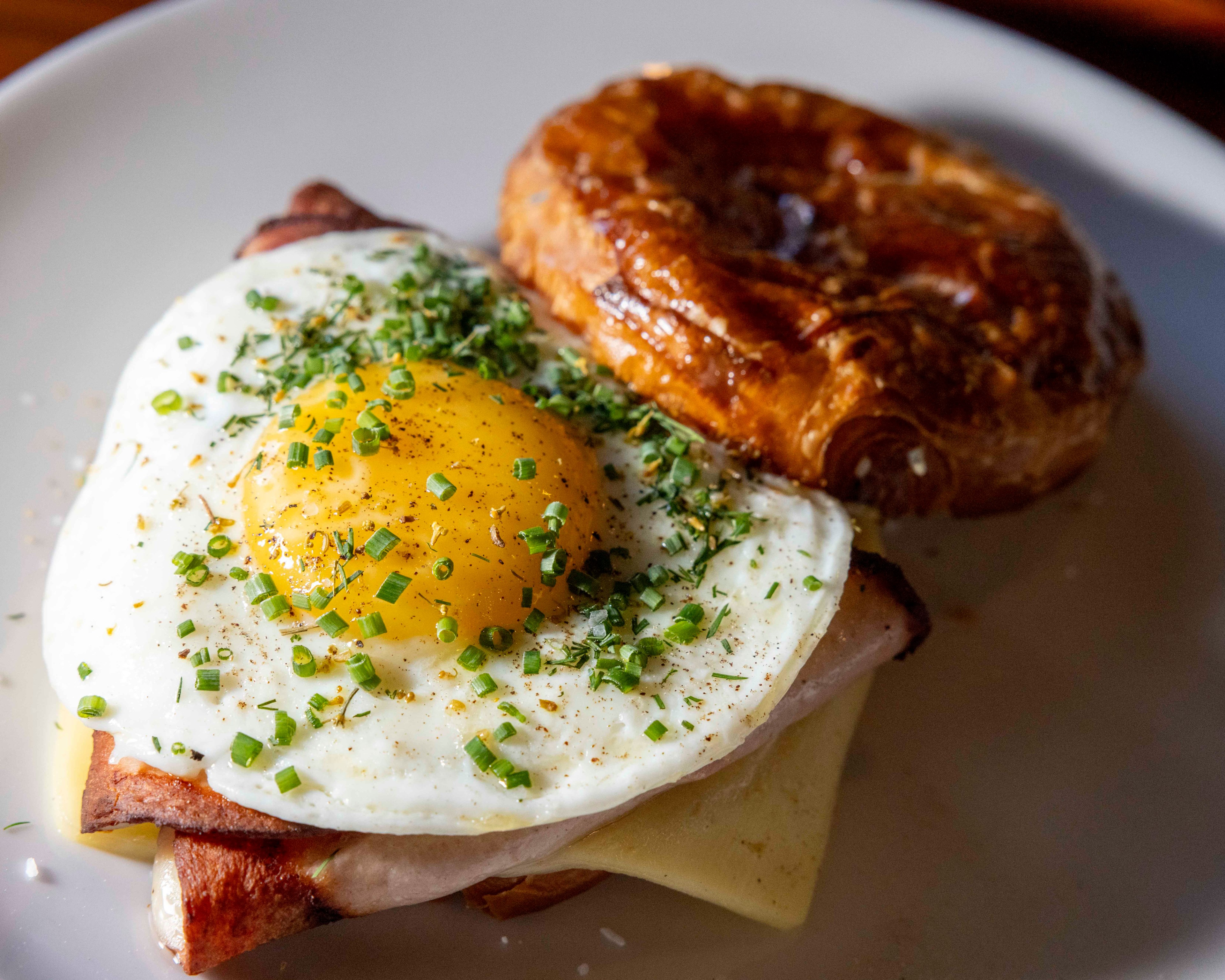 A breakfast sandwich features a sunny-side-up egg topped with green herbs, cheese, and ham on a flaky croissant, all presented on a white plate.