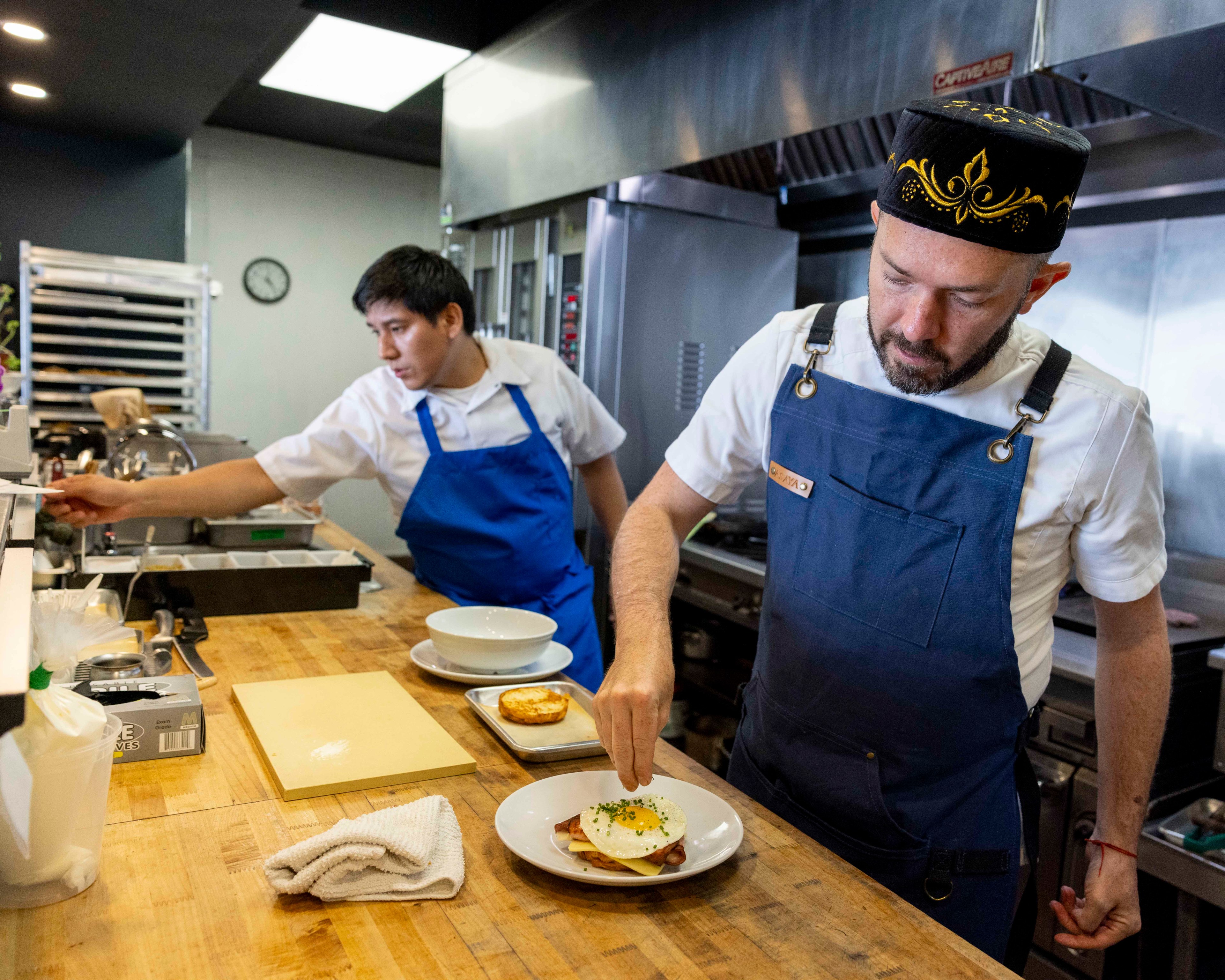 Two chefs in blue aprons work in a modern kitchen; one, wearing a decorated black hat, garnishes a dish; the other adjusts something, and various kitchen tools and ingredients are on the counter.