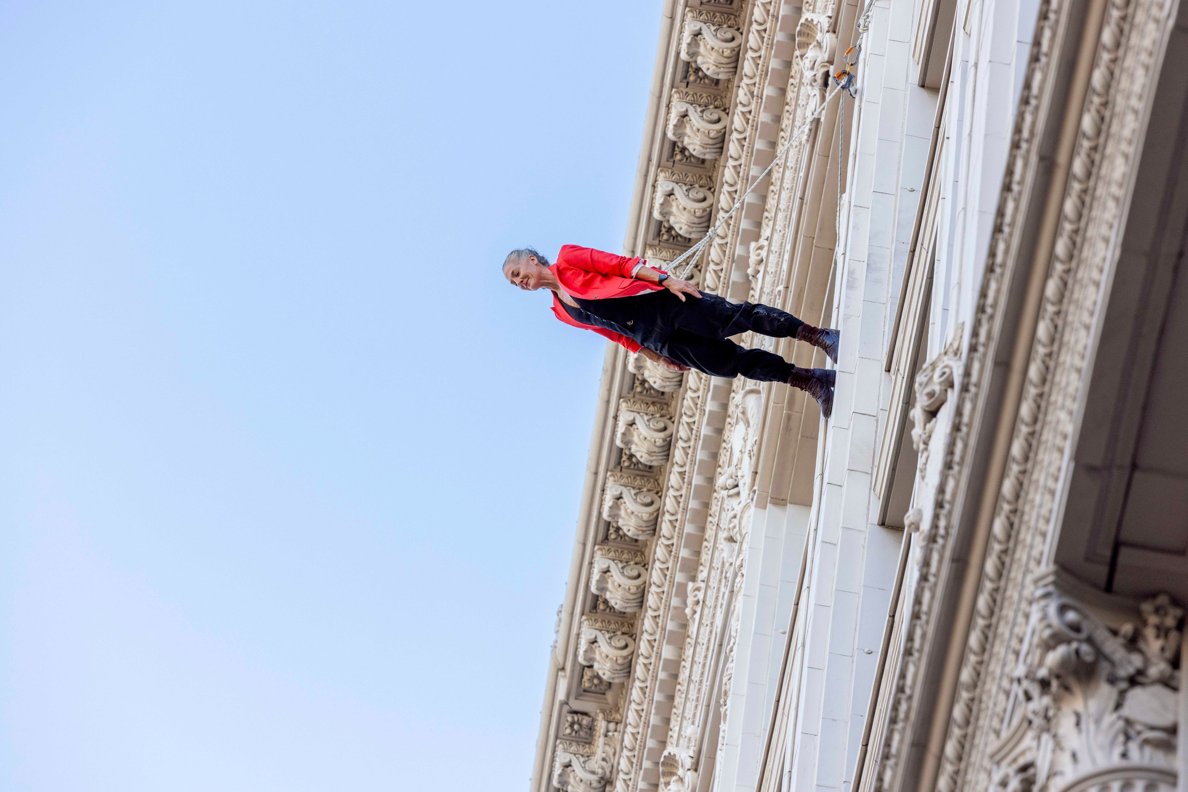 A person in a red jacket and black pants is suspended horizontally on the side of an ornate building, seemingly defying gravity against a clear blue sky.