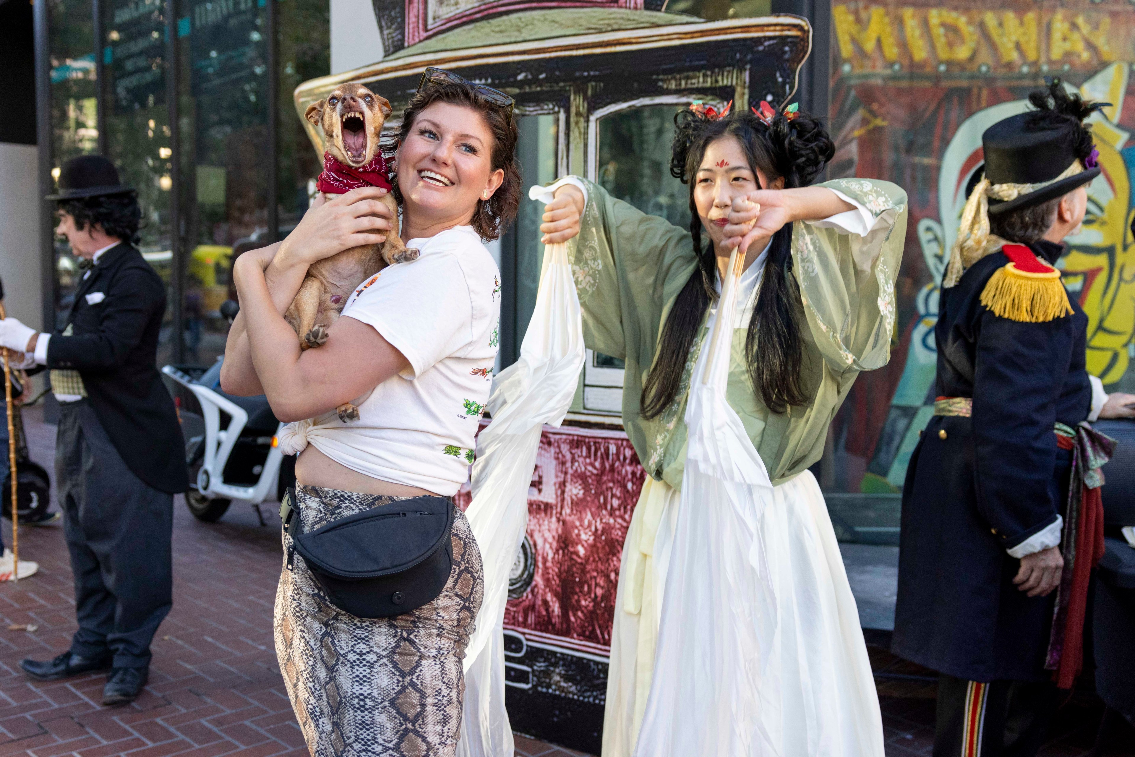 A smiling woman holds a small dog while another woman in traditional costume dances; people in vintage attire are seen in the background.