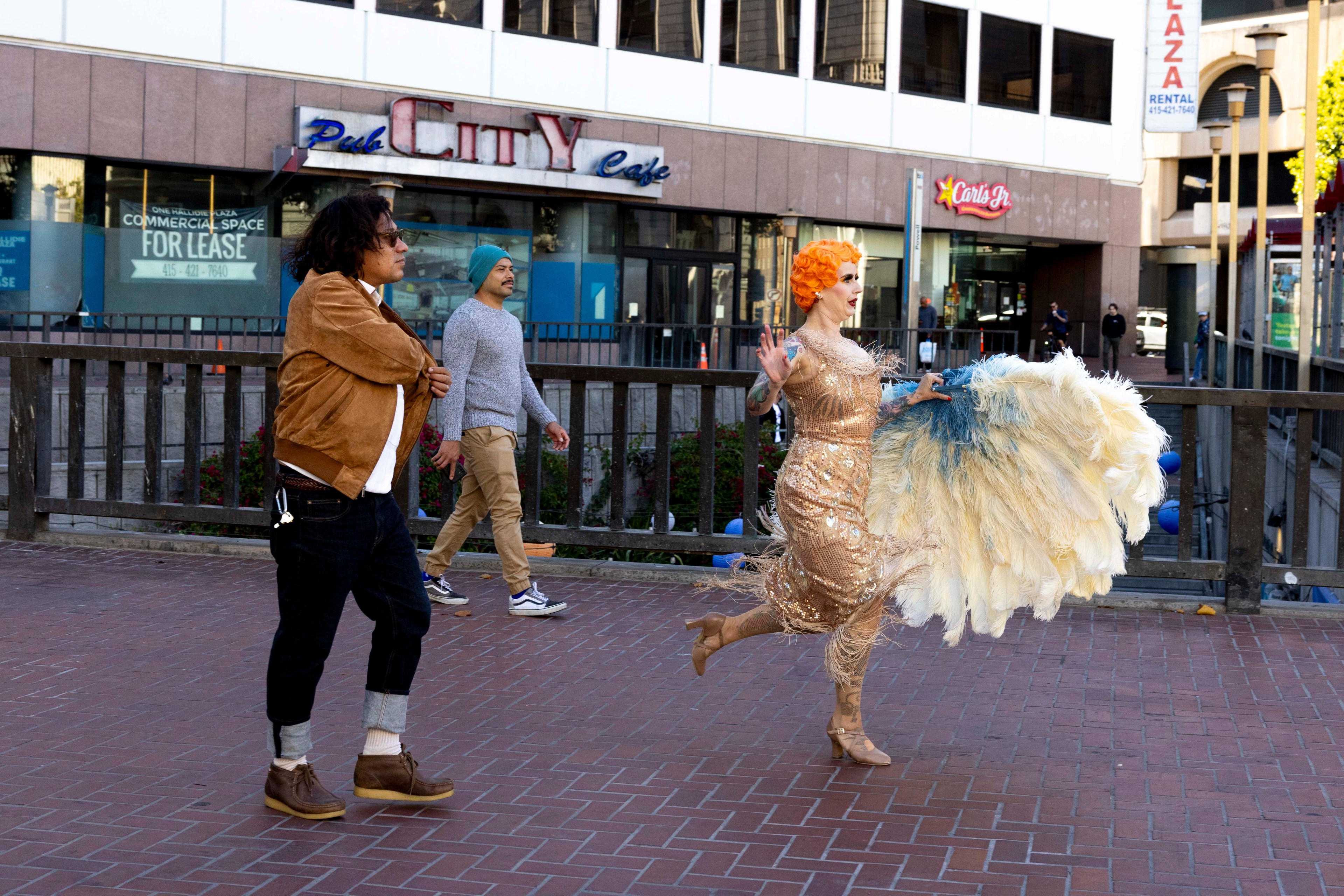 Three people are walking outdoors: one man in a brown jacket, another in a blue hat, and a woman in a sparkling dress carrying large, feathered fans.