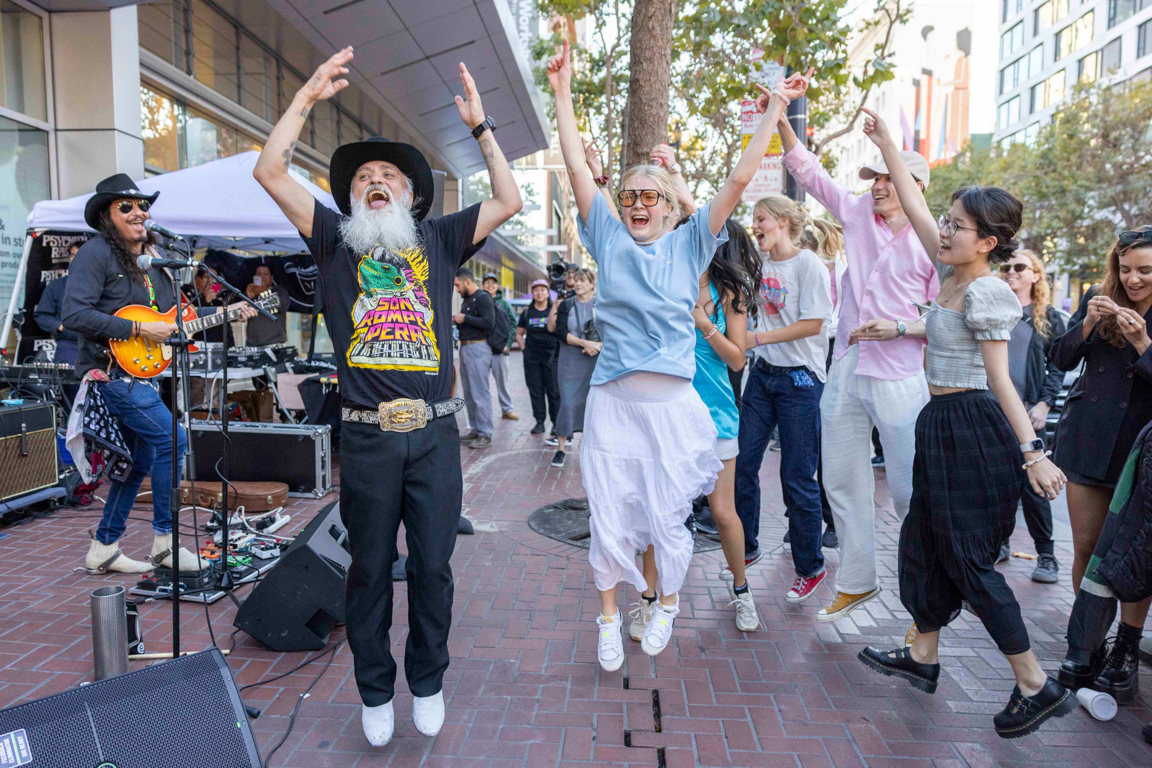 A lively street performance with a guitarist and a group of people joyfully dancing and jumping in unison, capturing a vibrant and energetic moment.