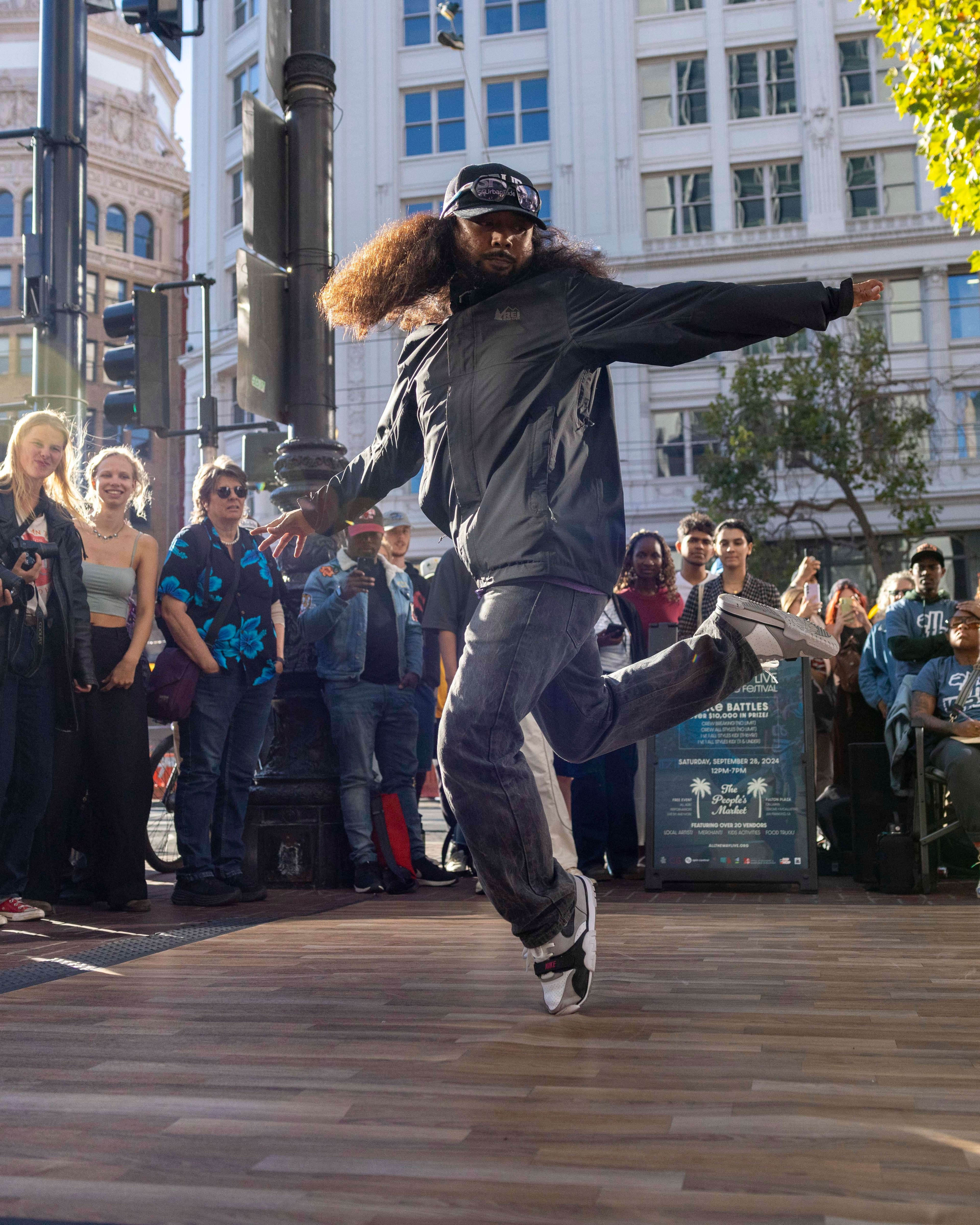 A street dancer with thick, curly hair performs energetically in front of an engaged crowd on a wooden platform in an urban area with tall buildings and trees.