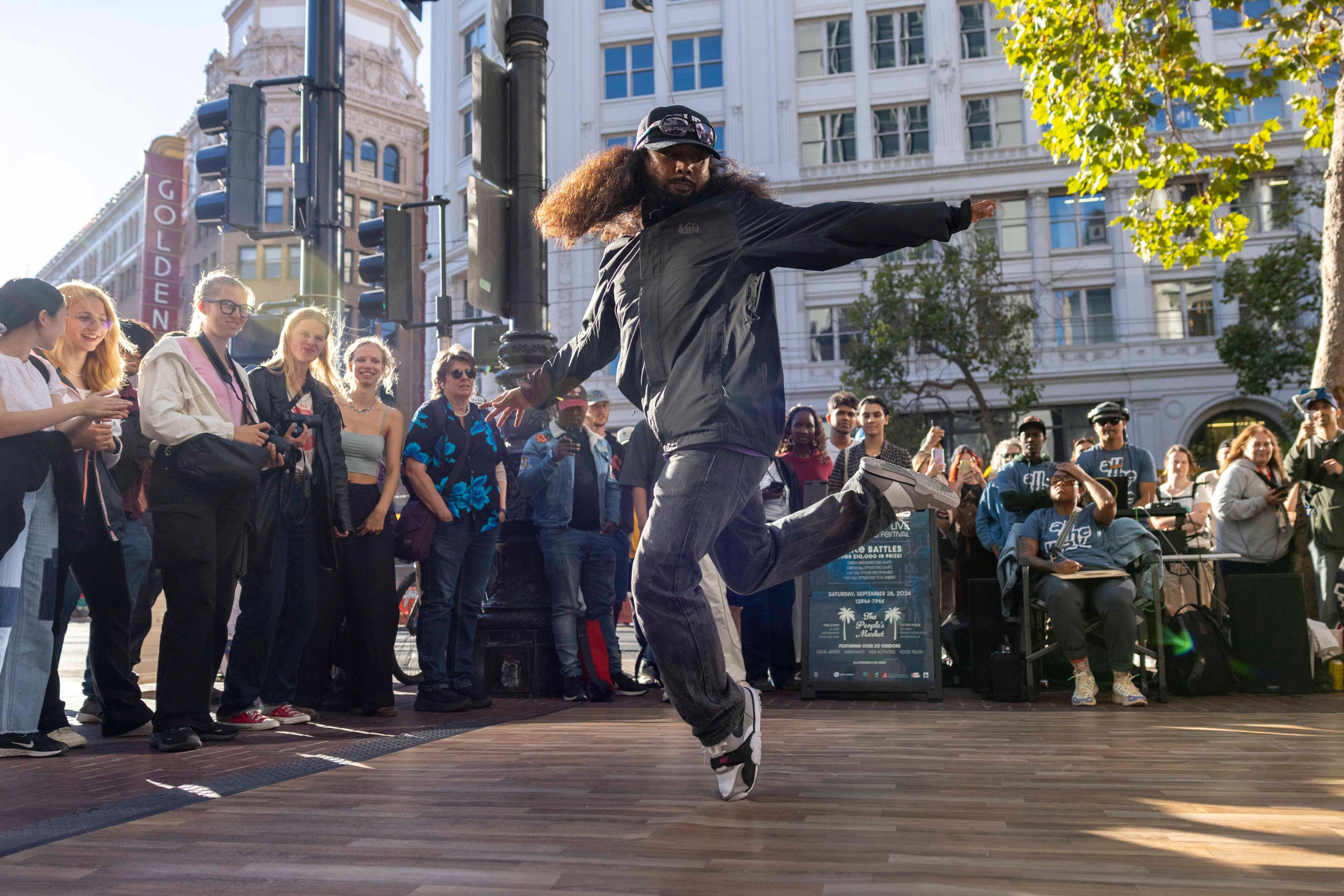 A man with long hair and a cap performs a dance move on a wooden platform in front of an engaged crowd on a city street, with buildings and trees in the background.