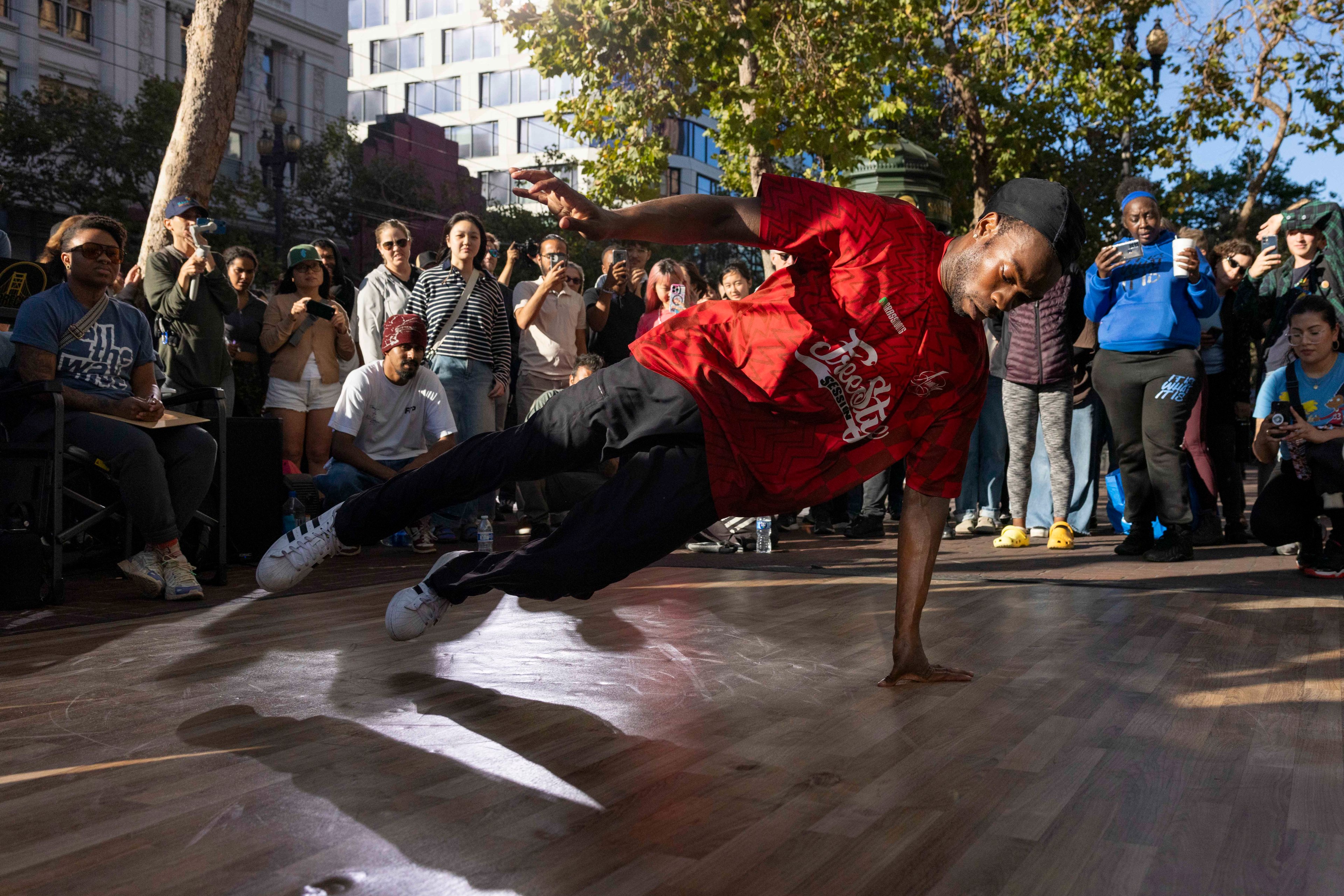 A street dancer in a red shirt performs a dynamic move on a wooden floor, while a crowd of onlookers, some filming, gathers around in an outdoor urban setting.