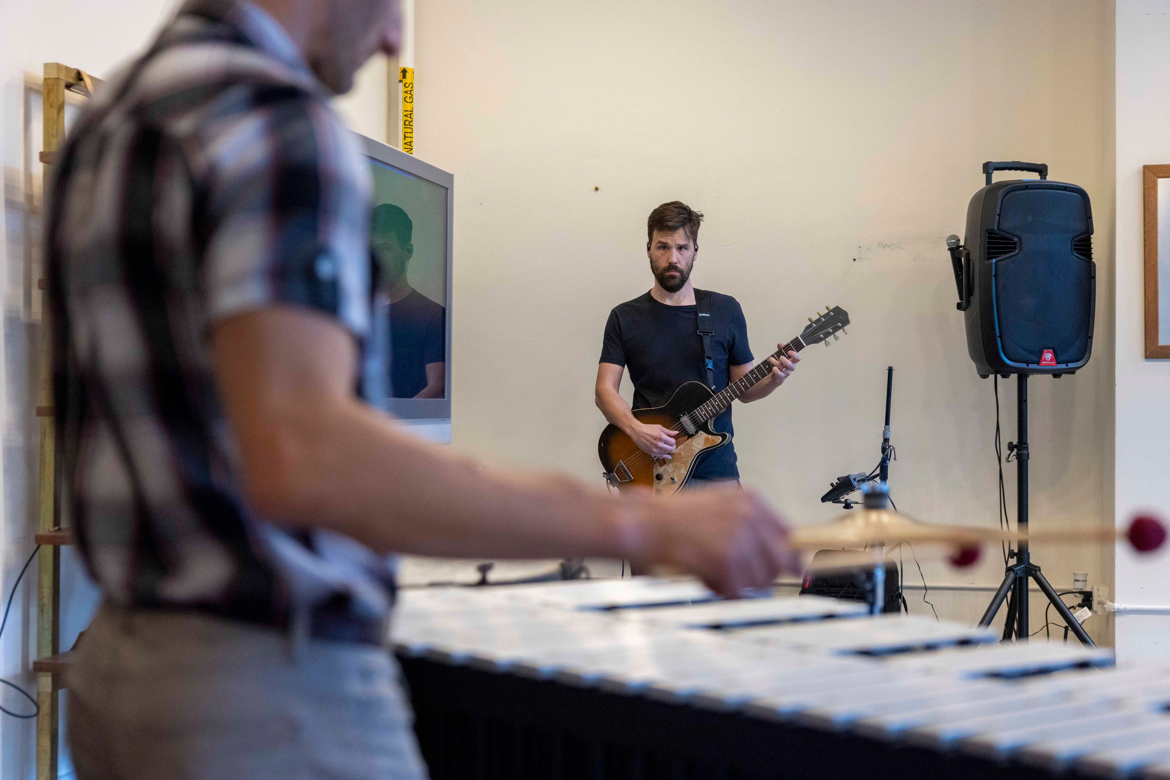 Two men are playing instruments in a room. One is focused on a vibraphone in the foreground, and the other is playing an electric guitar near a speaker in the background.