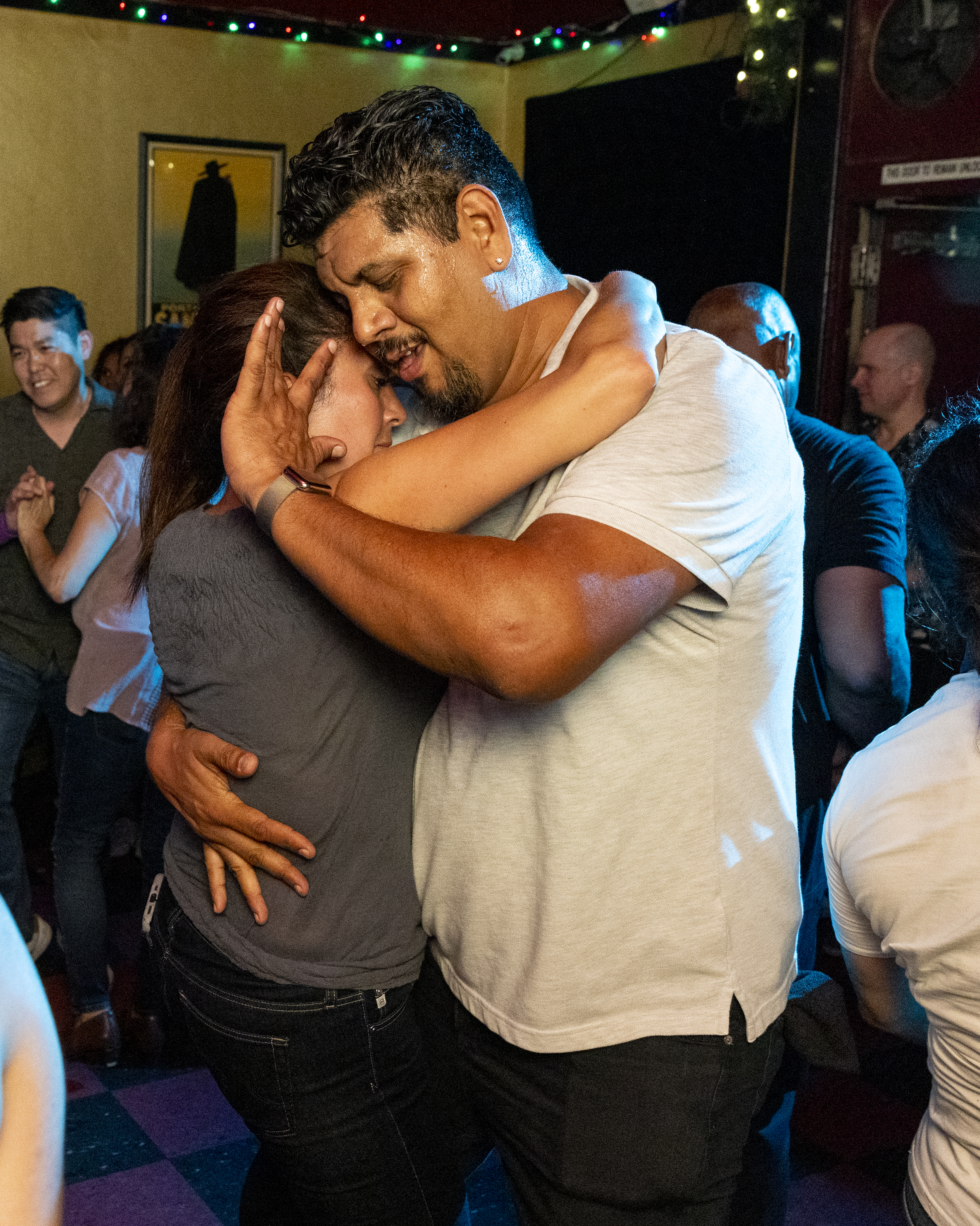 A couple is dancing closely with their eyes closed in a dimly lit room filled with other dancing people. The mood is intimate and joyful.