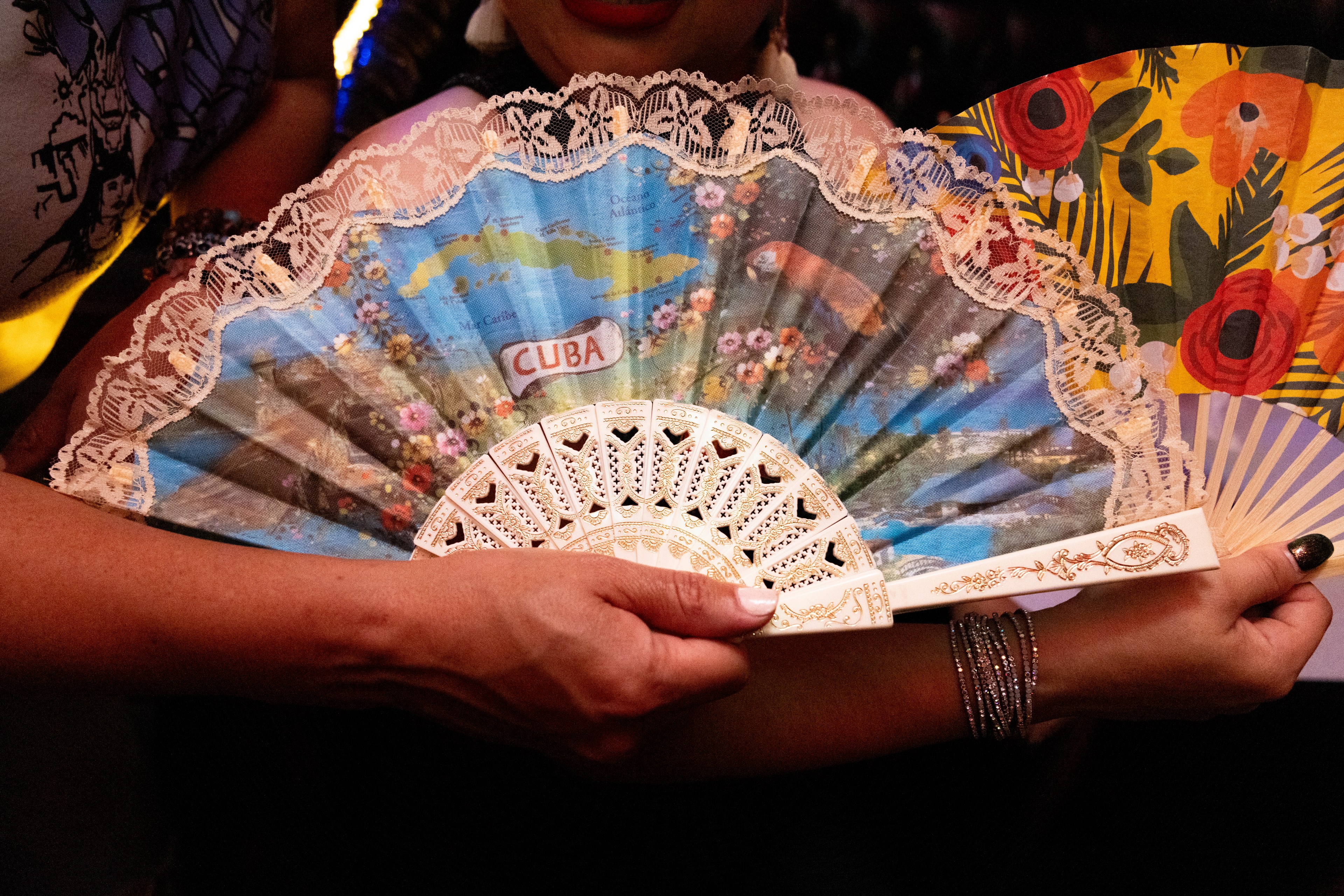 A person holds an intricately designed fan with a colorful map of Cuba and floral patterns, framed with lace. Behind it is another fan with a vibrant floral design.