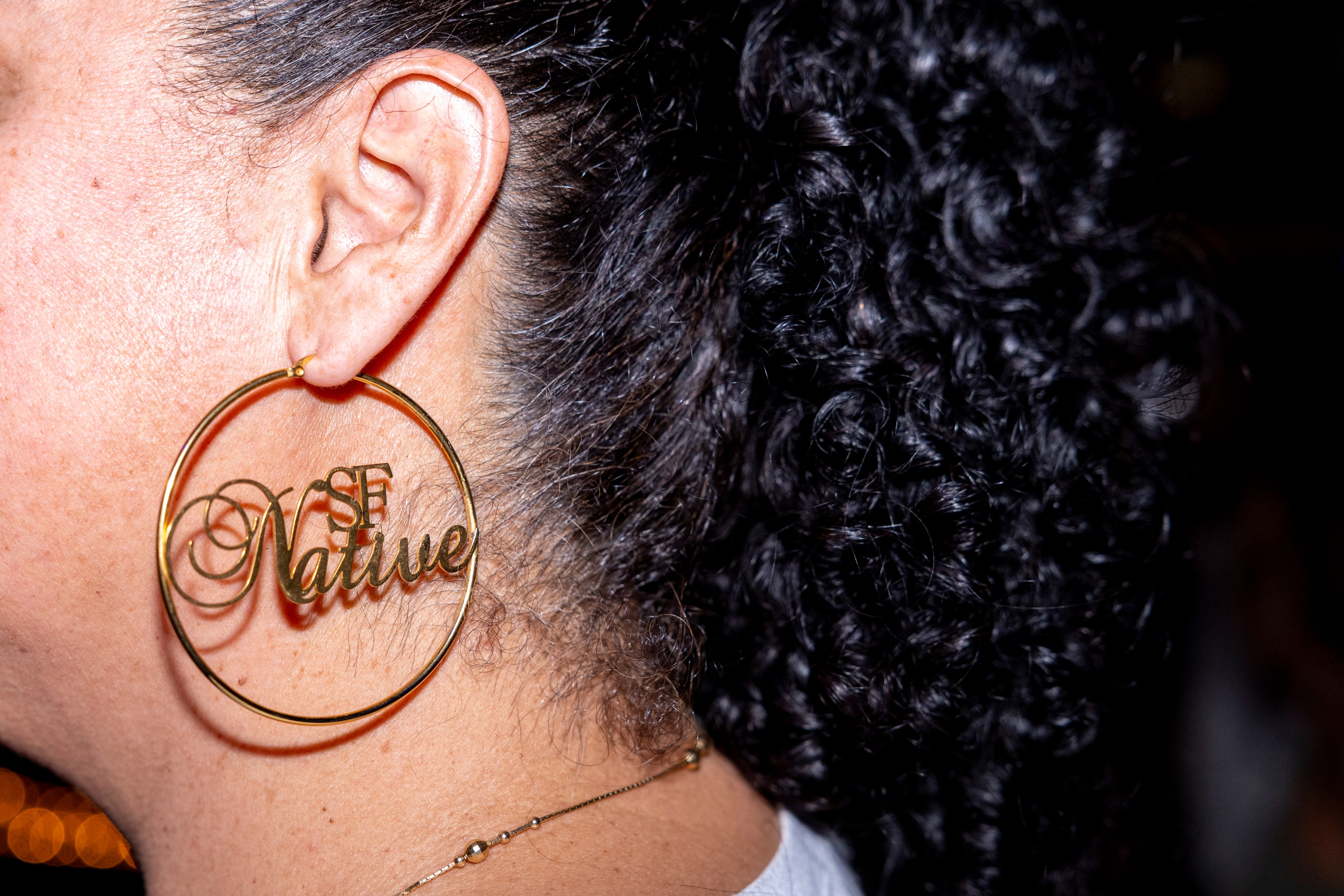 The image shows a close-up of a person's ear with a large, gold hoop earring that reads &quot;SF Native&quot; in a cursive font. The person has curly dark hair.