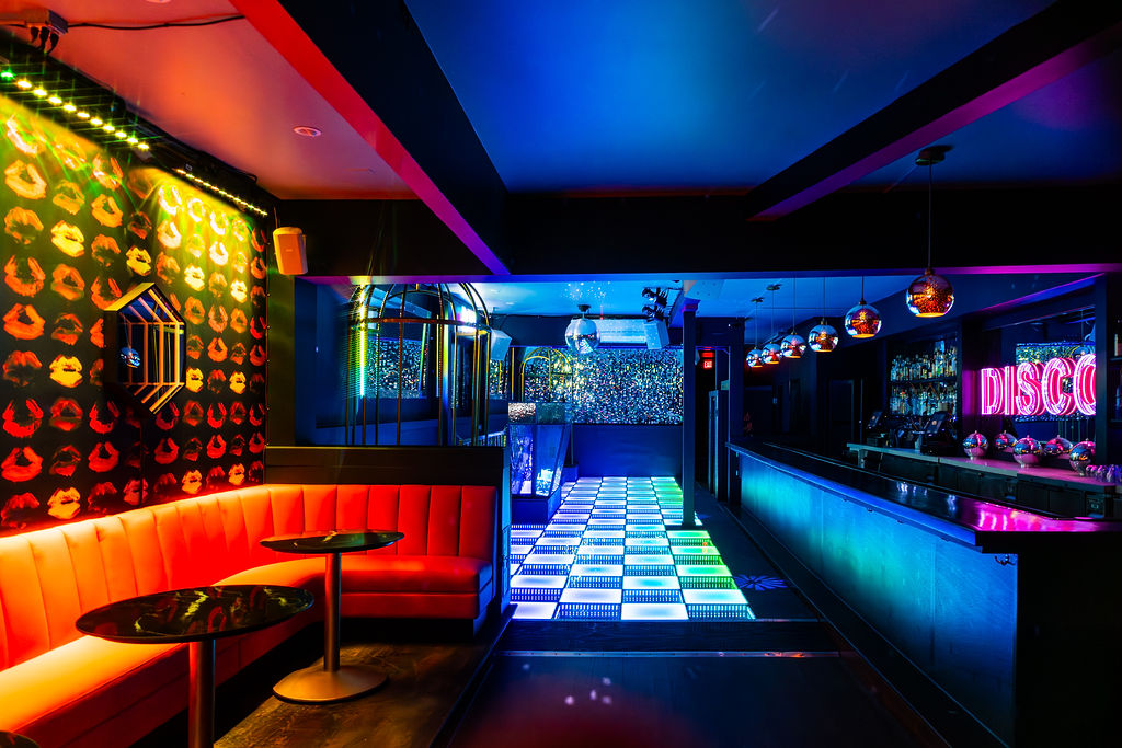 A vibrant, colorful nightclub with a light-up dance floor, neon &quot;DISCO&quot; sign, red seating, walls adorned with lip prints, and a long, illuminated bar.