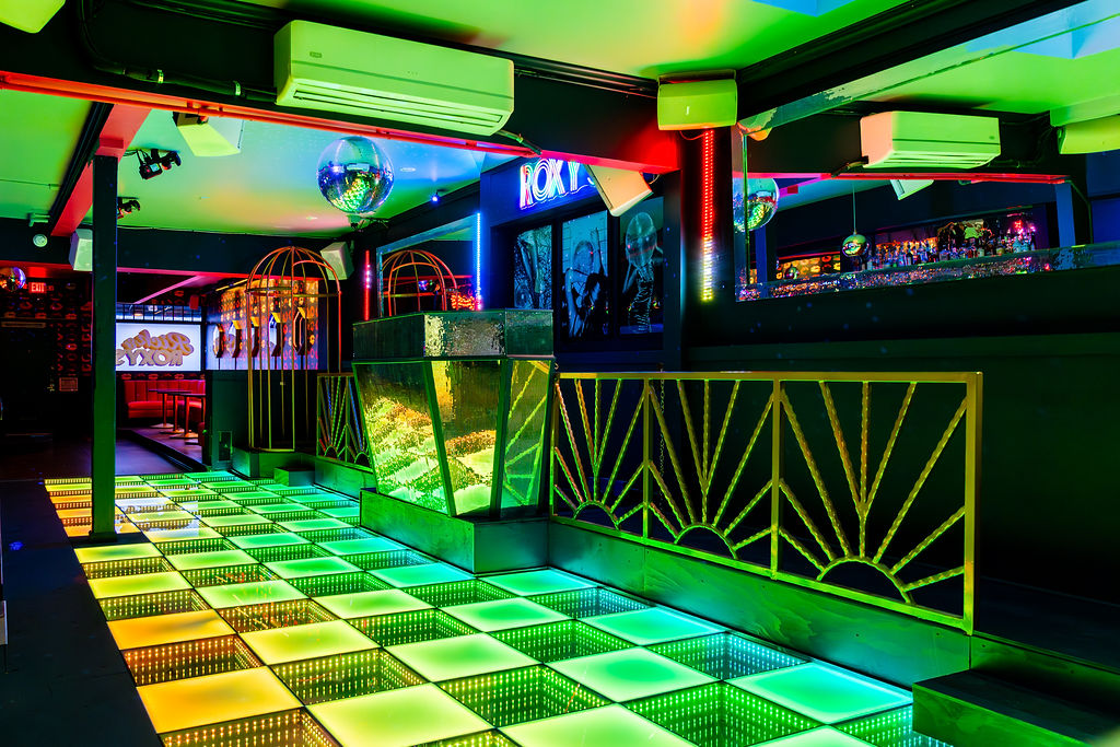 A nightclub with a neon-lit dance floor, disco balls, and futuristic decor featuring mirrors, golden railings, and colorful lighting accents throughout the space.