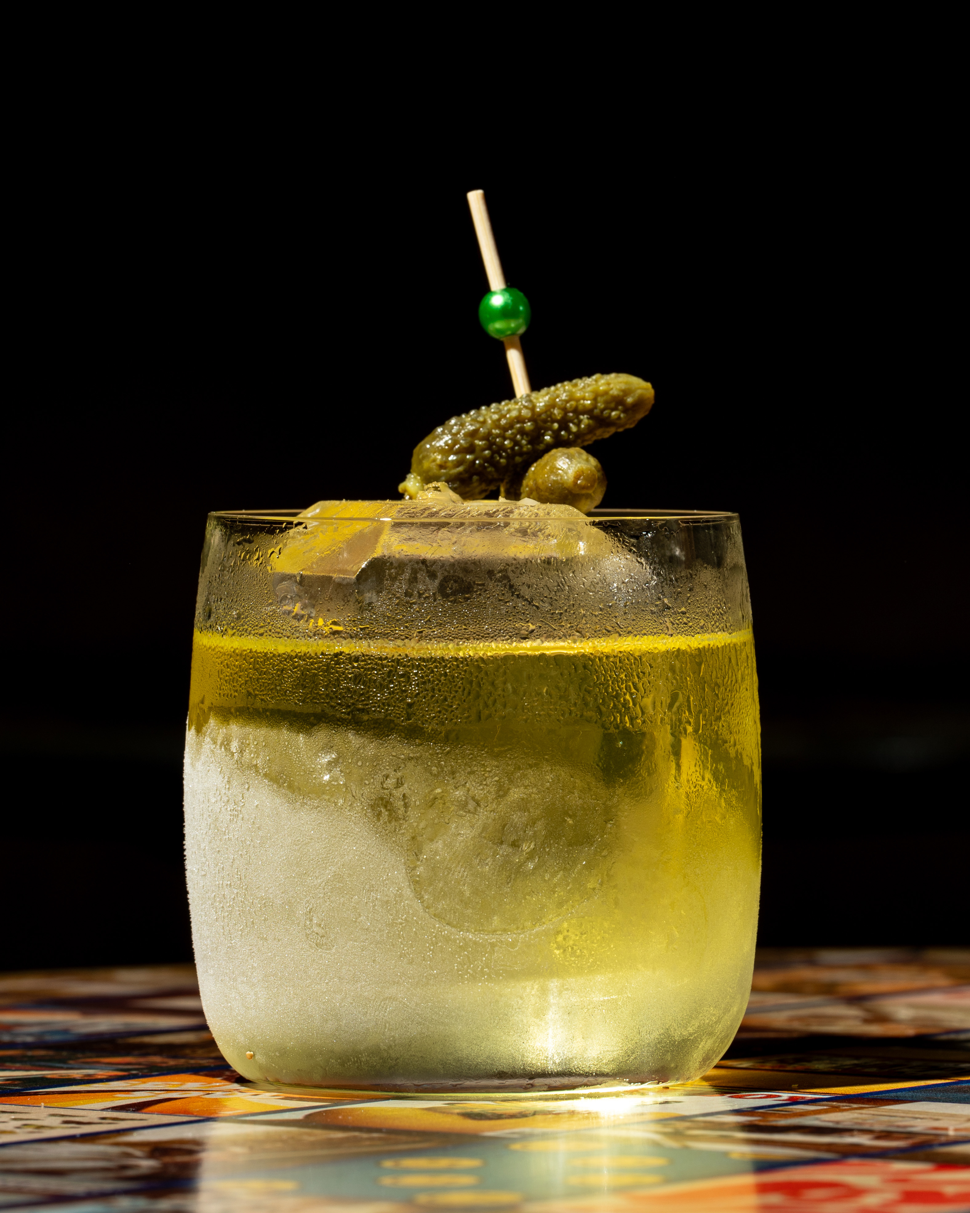 A cocktail glass filled with a yellowish liquid and ice is garnished with a mini pickle and an olive on a skewer, set against a dark background. 