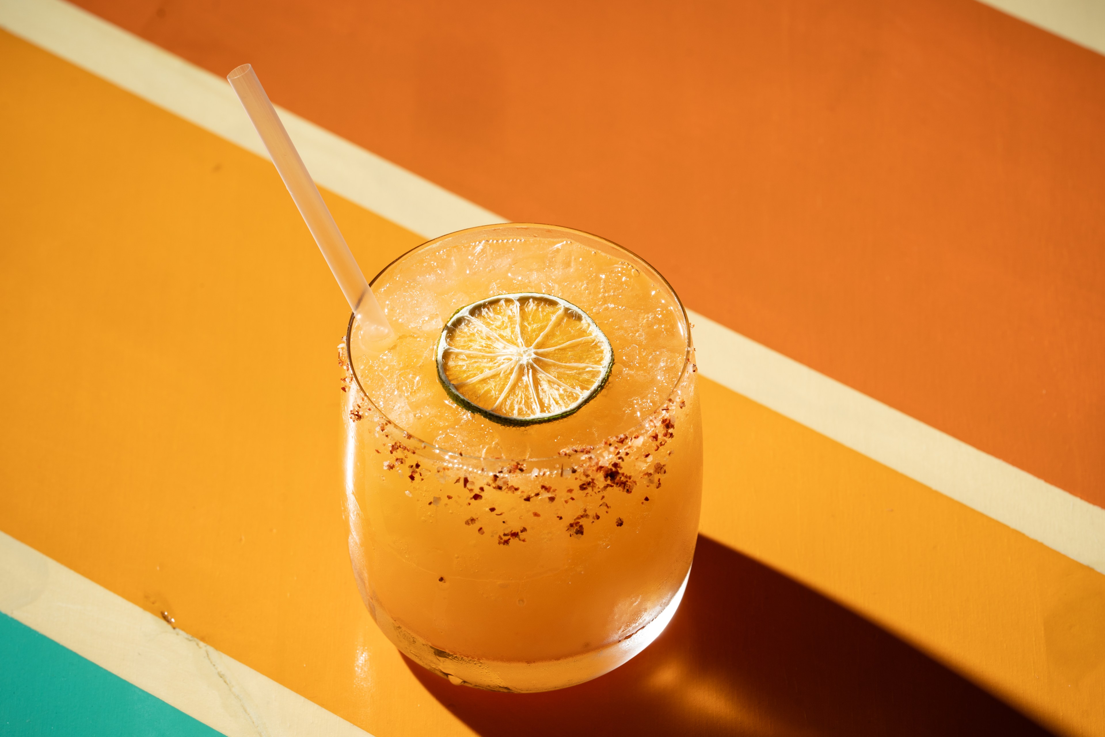 A vibrant orange drink with ice is served in a glass with a slice of lime and a straw, set against a background of orange and yellow diagonal stripes.