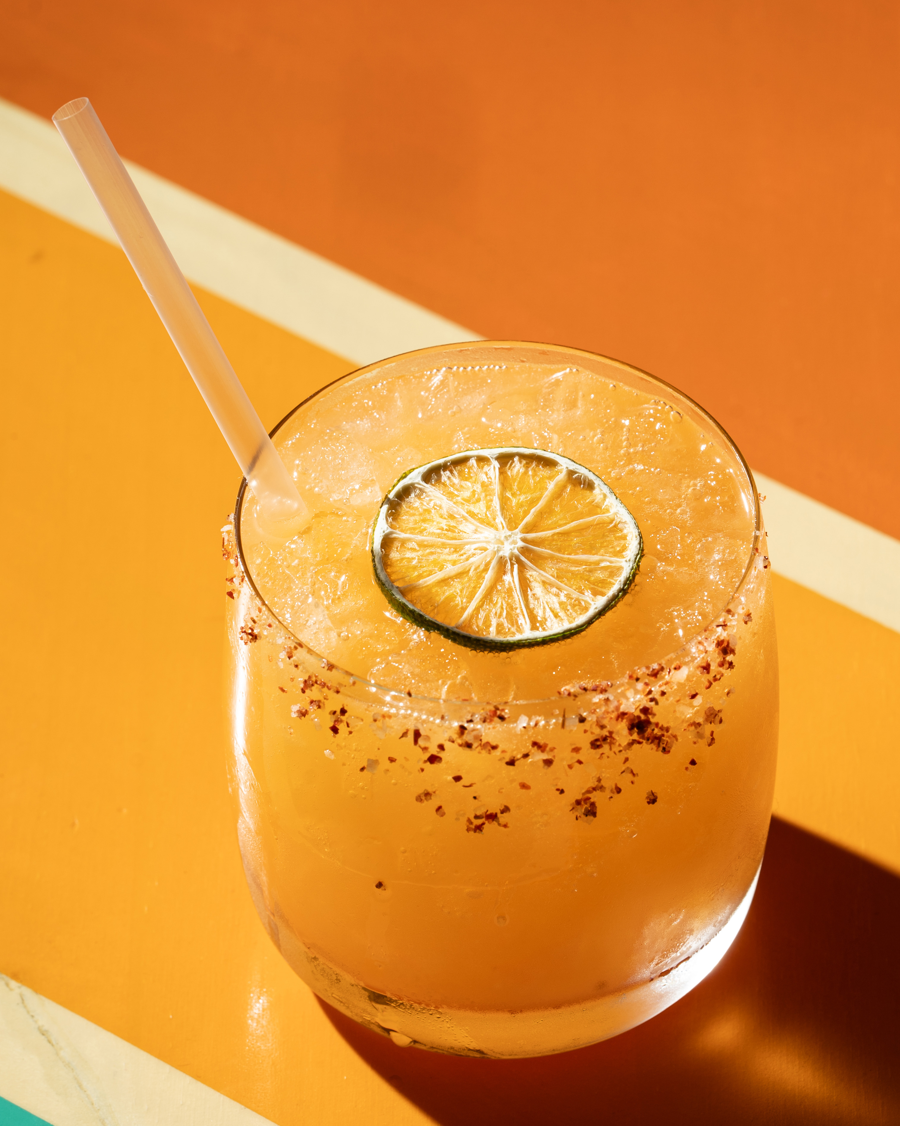 A refreshing orange drink garnished with a lime slice and a chili powder-rimmed edge sits on an orange and yellow surface, featuring a straw.