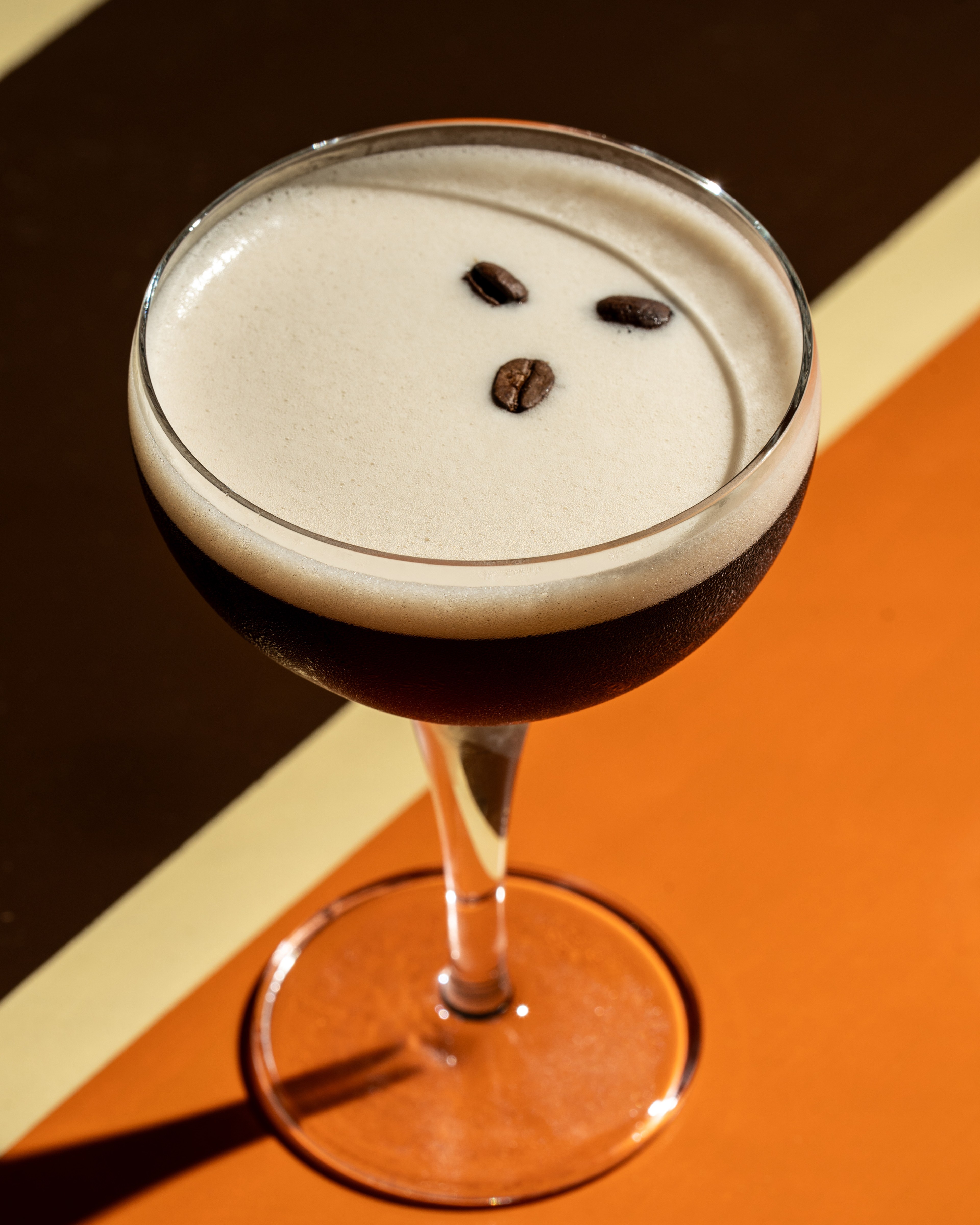 A frothy espresso martini in a cocktail glass is garnished with three coffee beans, set against a stylish, striped brown and orange backdrop.