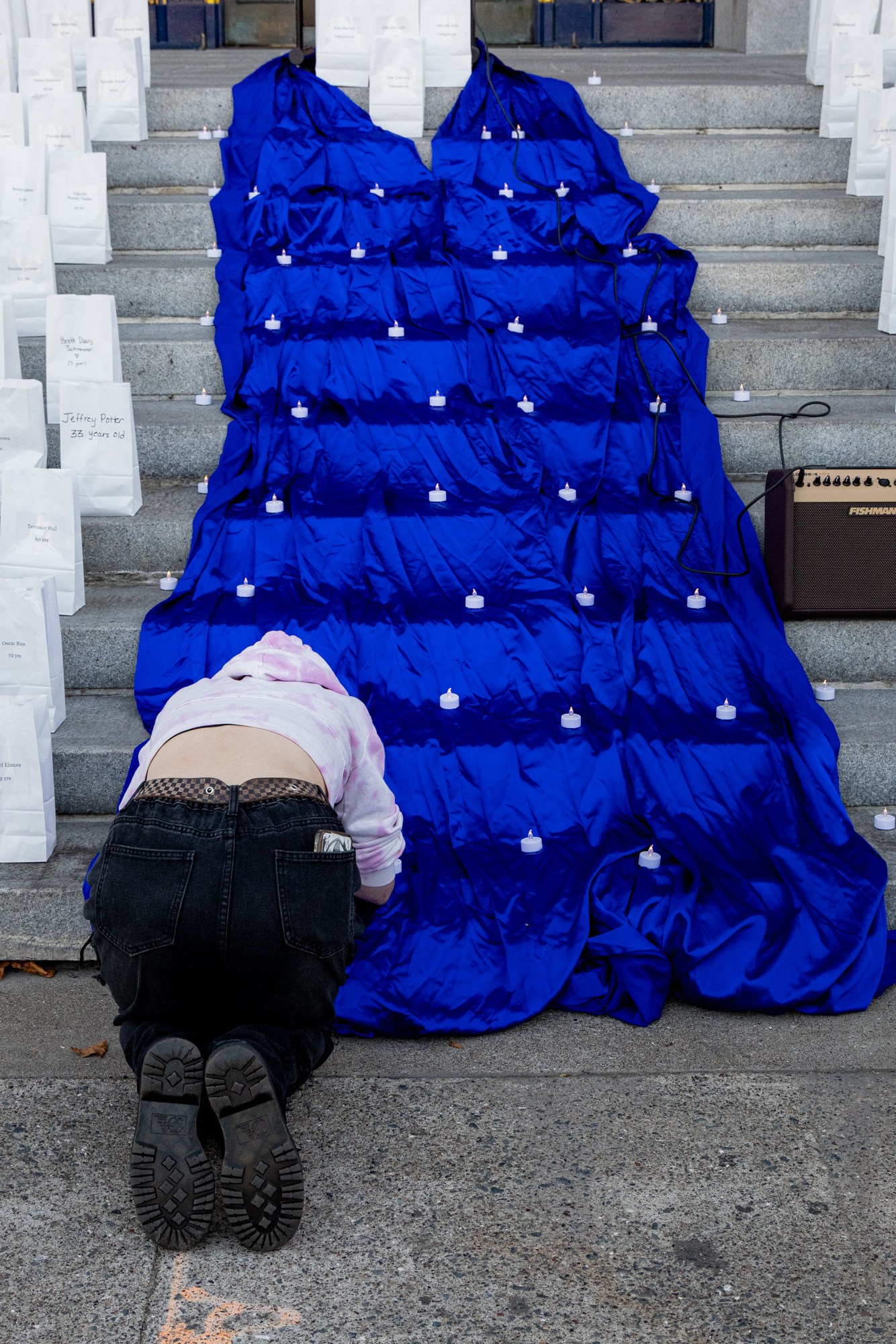 A person kneels on a staircase covered in a blue cloth with small lights. Surrounding them are numerous white bags, each labeled, and a speaker sits on the steps.