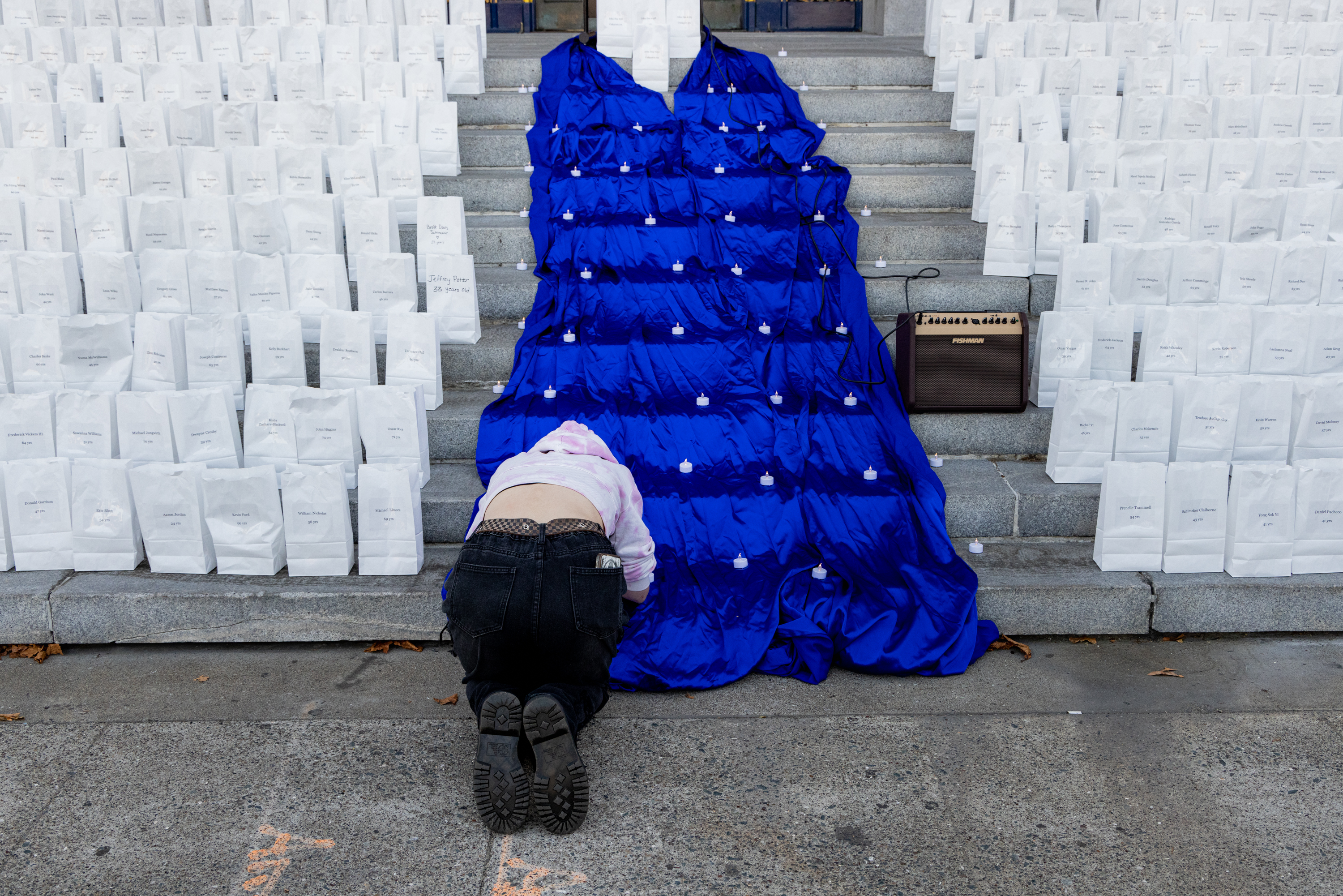 A person kneels on a staircase covered in a blue cloth with small lights. Surrounding them are numerous white bags, each labeled, and a speaker sits on the steps.