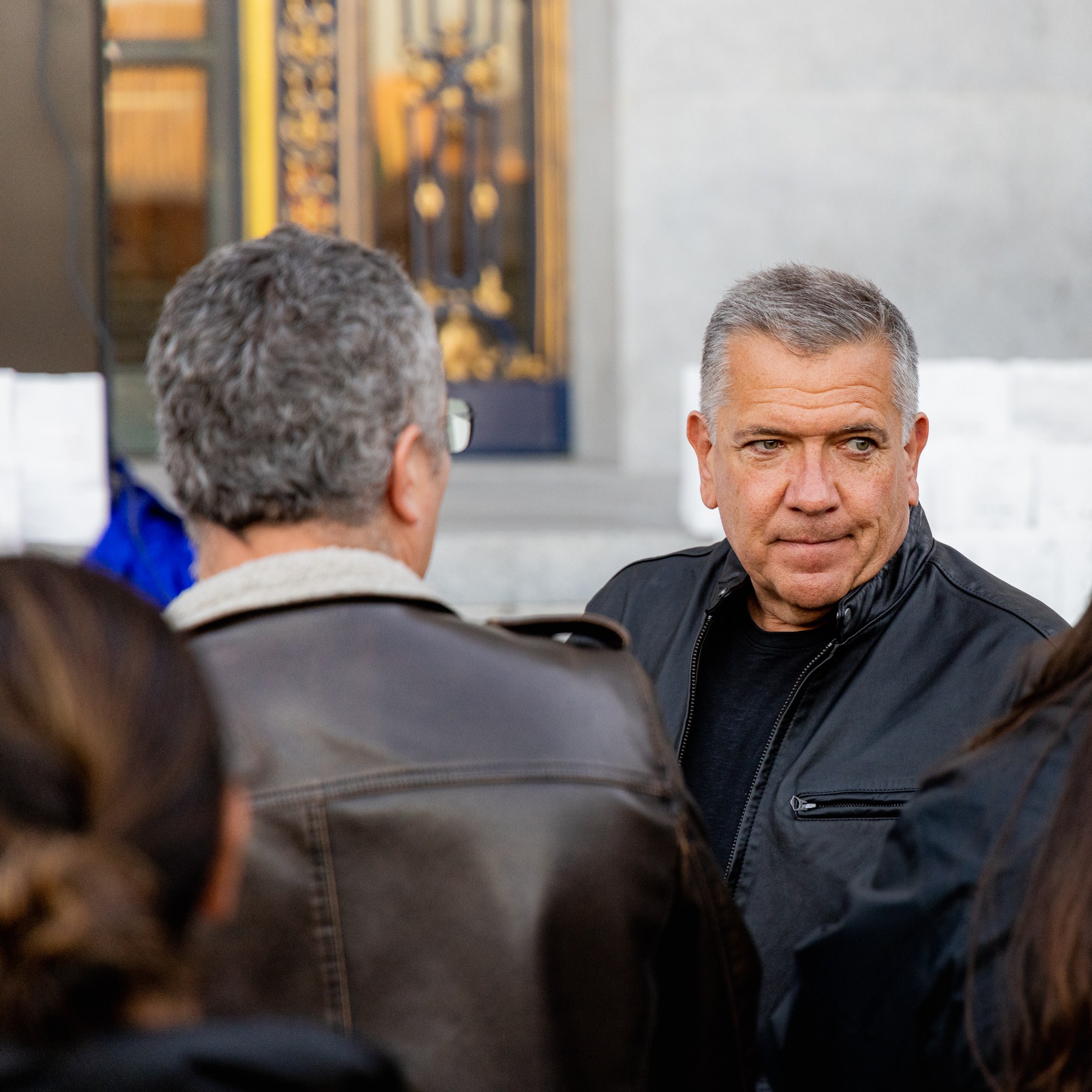 A man in a black jacket stands in a group, looking slightly to his right. Other people around him have their backs to the camera, and a decorative background is visible.