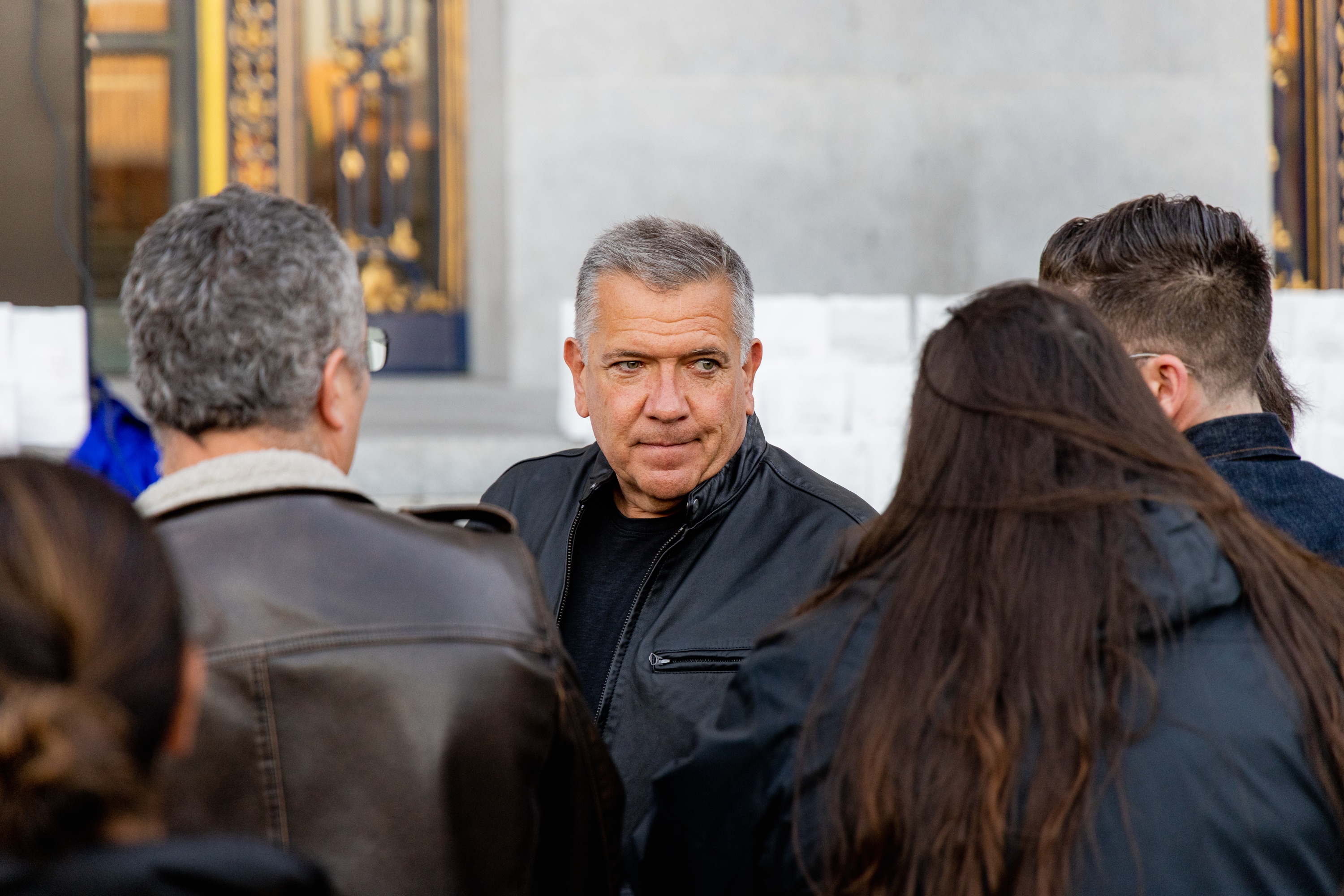 A man in a black jacket stands in a group, looking slightly to his right. Other people around him have their backs to the camera, and a decorative background is visible.