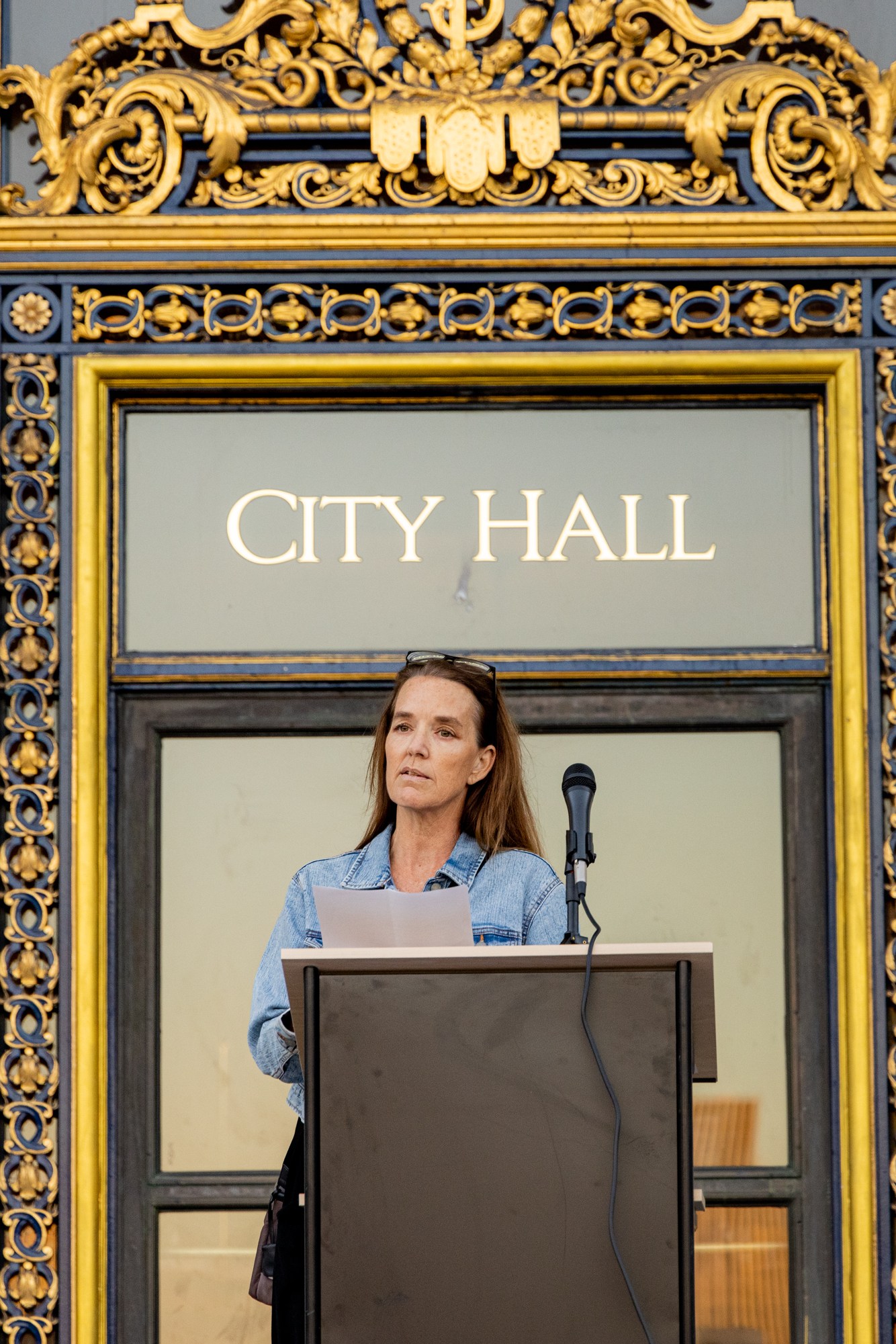 A woman stands behind a podium with a microphone in front of an ornate City Hall entrance, adorned with gold and intricate designs.