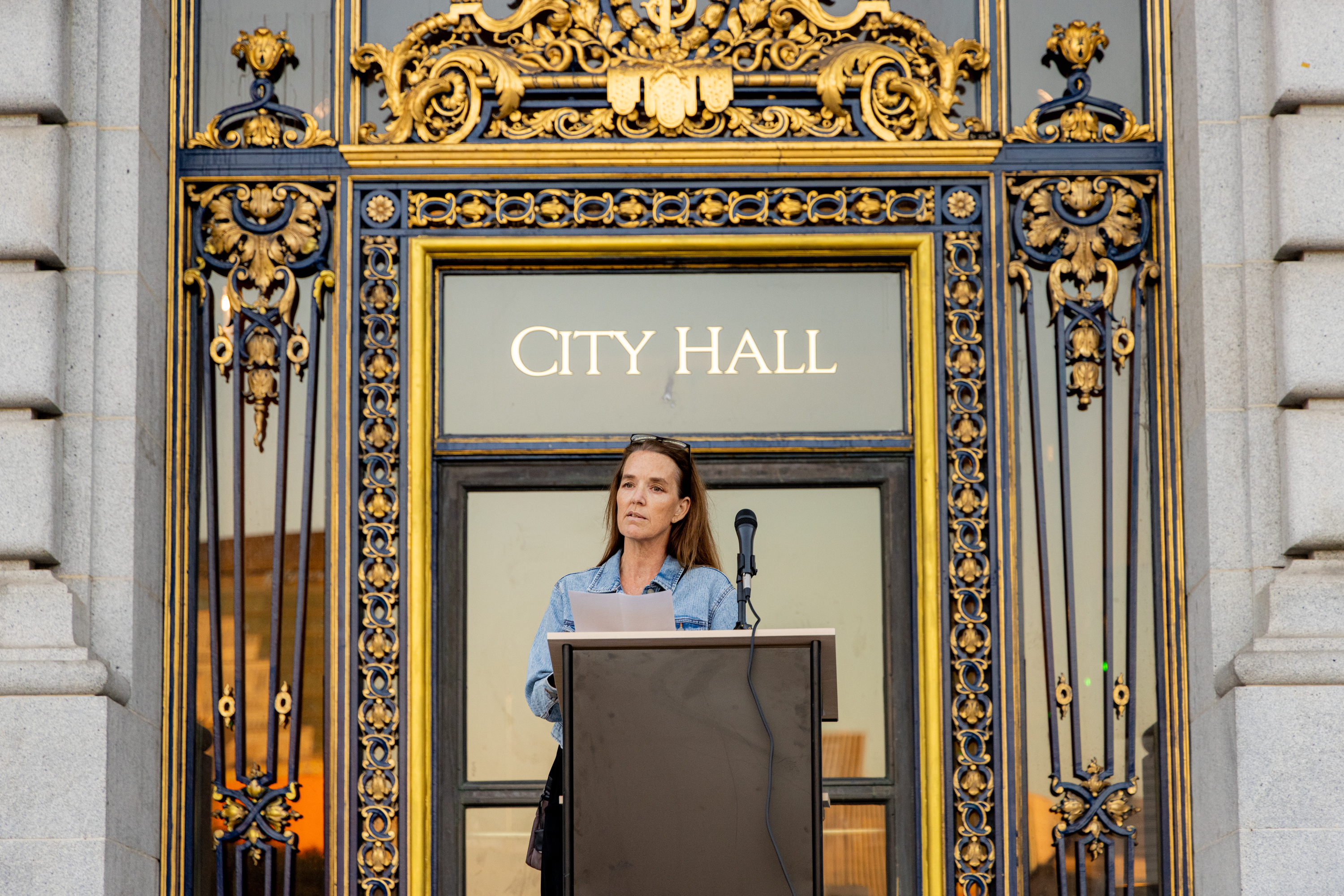 A woman stands behind a podium with a microphone in front of an ornate City Hall entrance, adorned with gold and intricate designs.