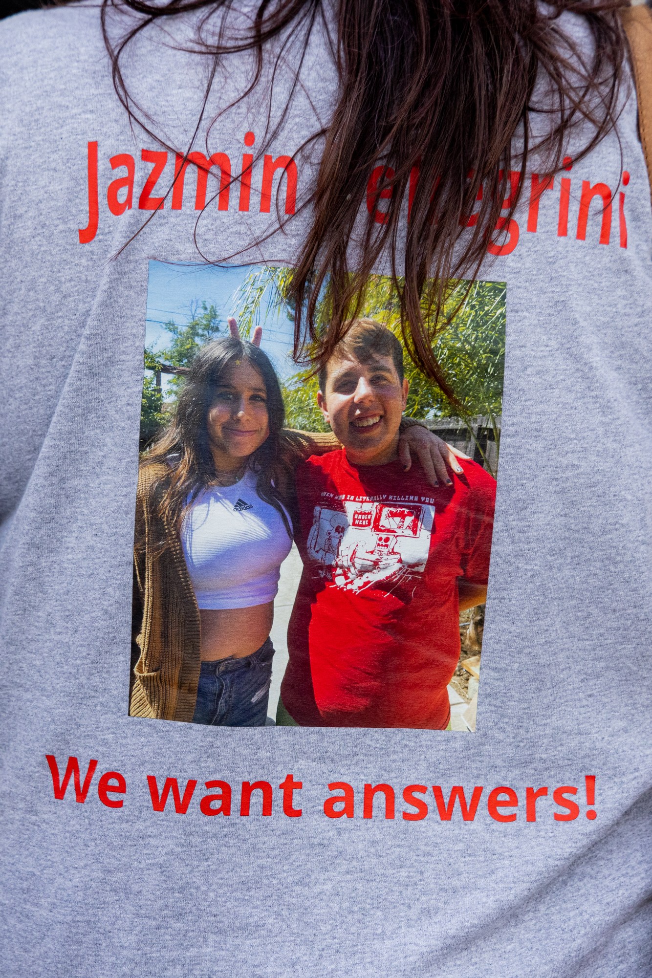 A person is wearing a gray t-shirt featuring a photo of two smiling individuals, with red text above saying &quot;Jazmin Pellegrini&quot; and below saying &quot;We want answers!&quot;