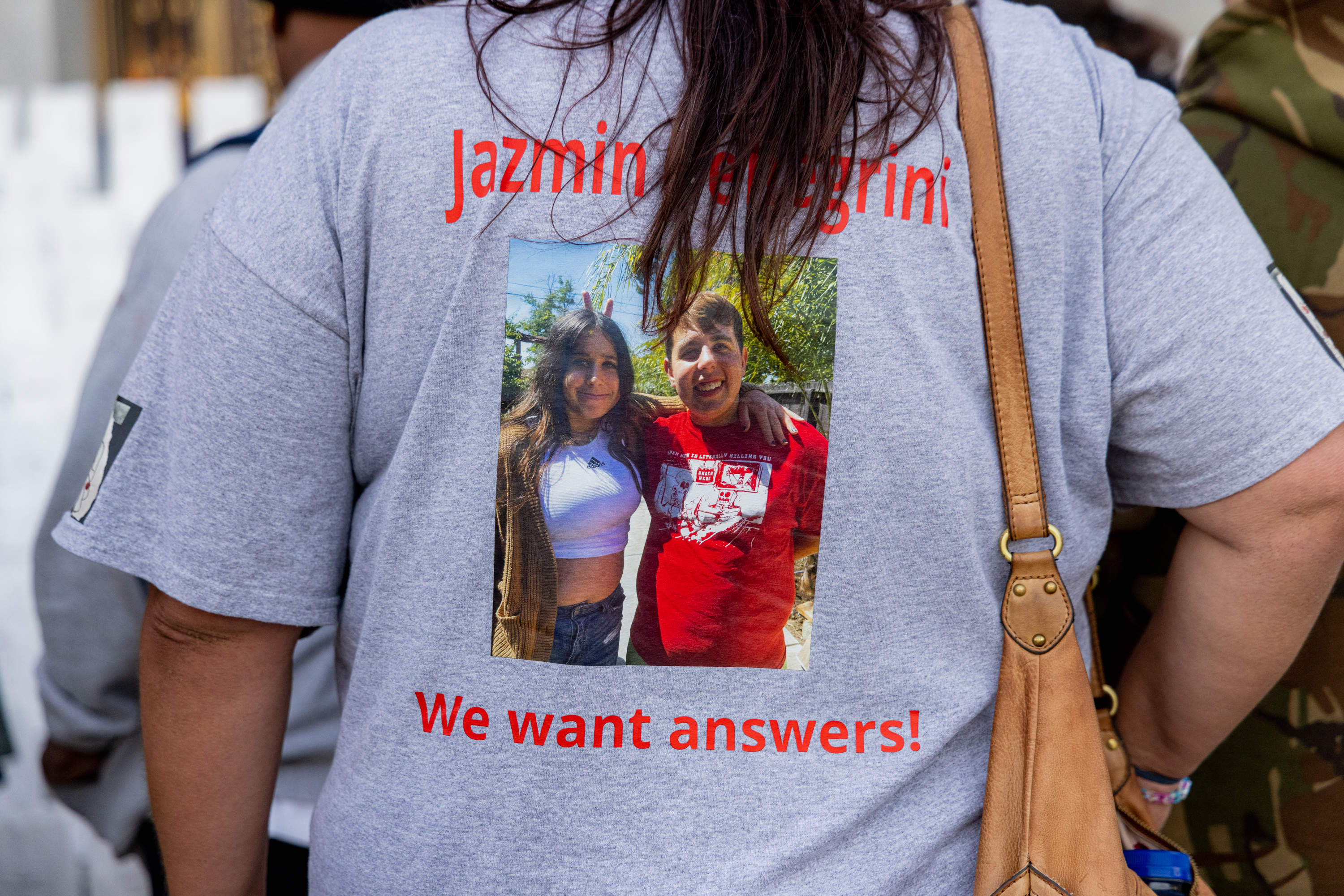 A person is wearing a gray t-shirt featuring a photo of two smiling individuals, with red text above saying &quot;Jazmin Pellegrini&quot; and below saying &quot;We want answers!&quot;