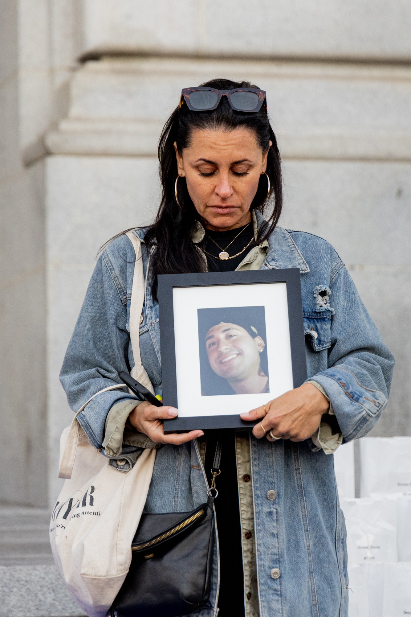 A woman stands solemnly, holding a framed photo of a smiling person. She wears a denim jacket, with sunglasses atop her head, in front of ornate doors with gold detailing.