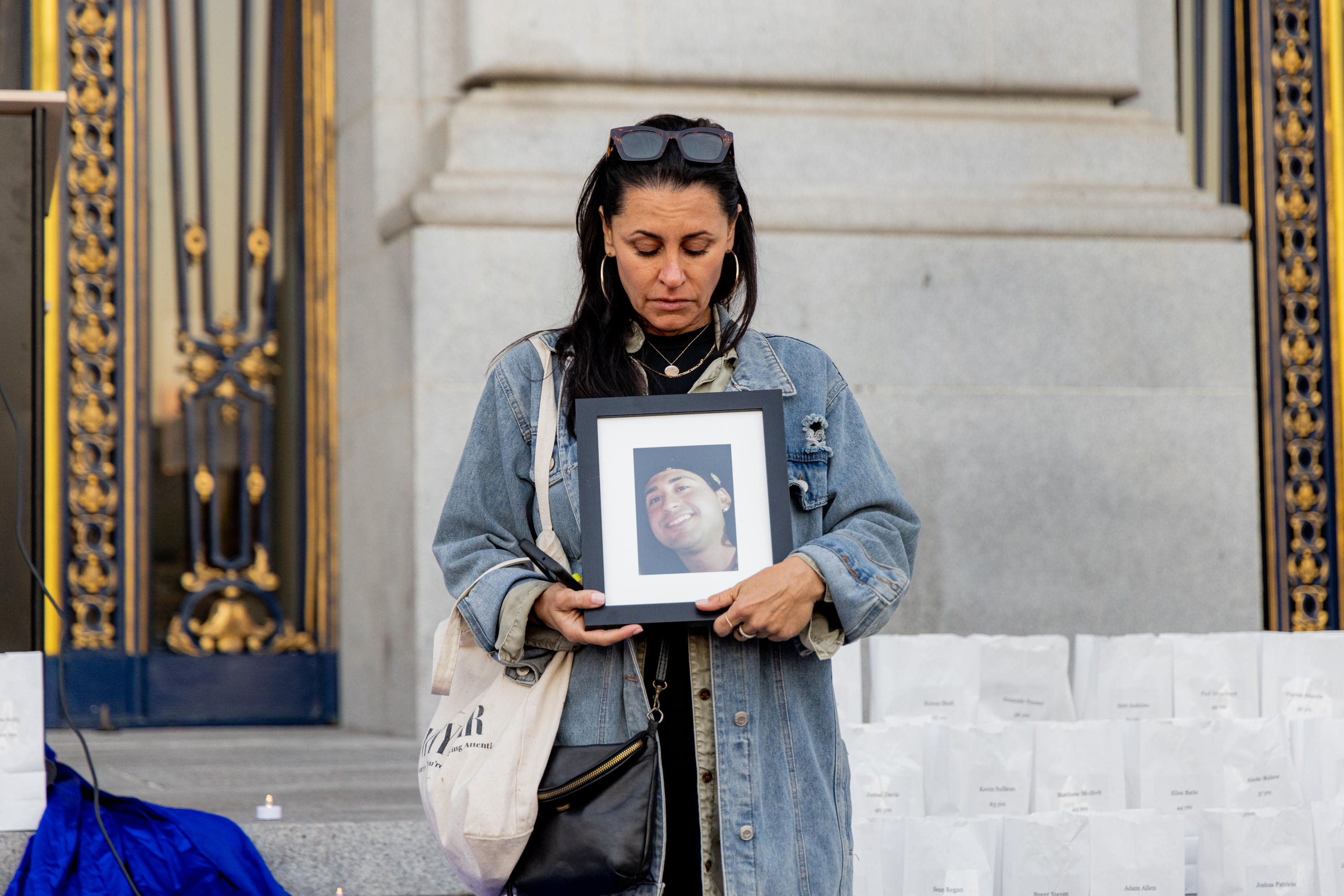 A woman stands solemnly, holding a framed photo of a smiling person. She wears a denim jacket, with sunglasses atop her head, in front of ornate doors with gold detailing.