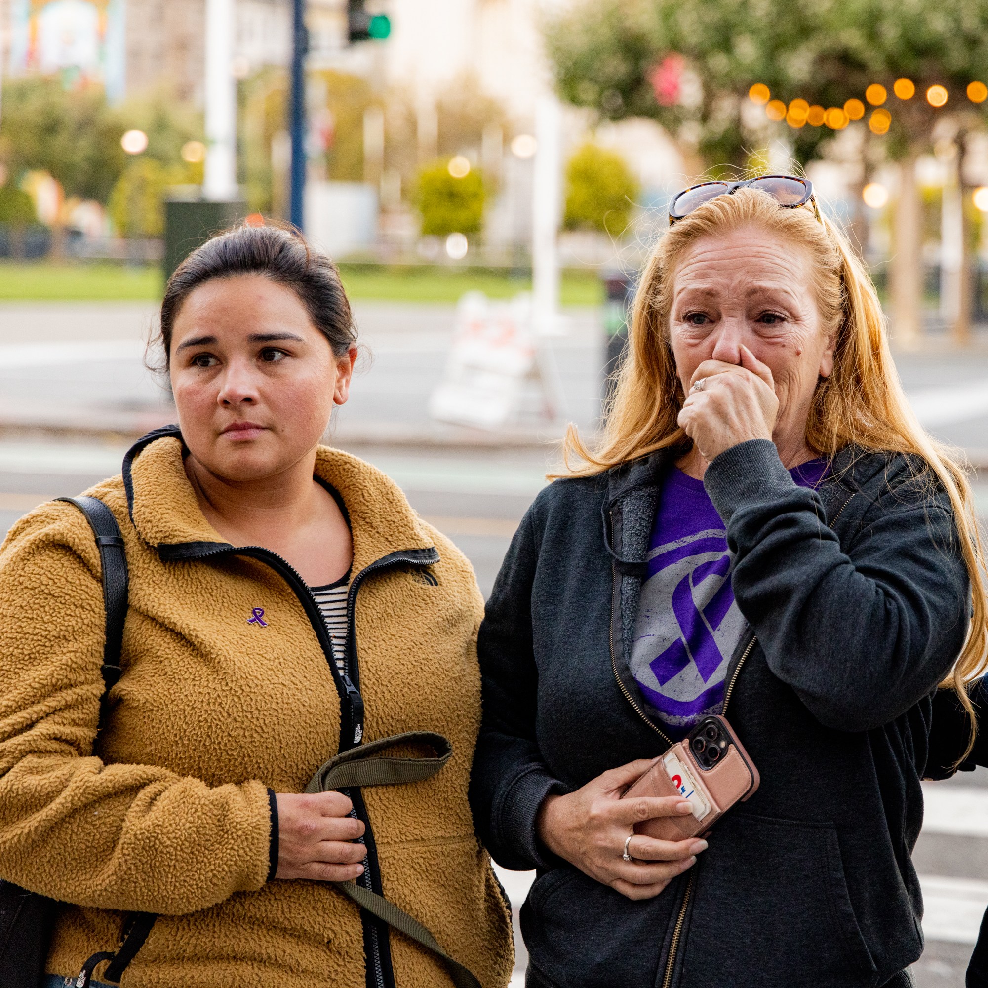 Three people stand outdoors; a woman wipes tears, clutching a phone, flanked by a serious-looking woman and a man looking down at his phone.