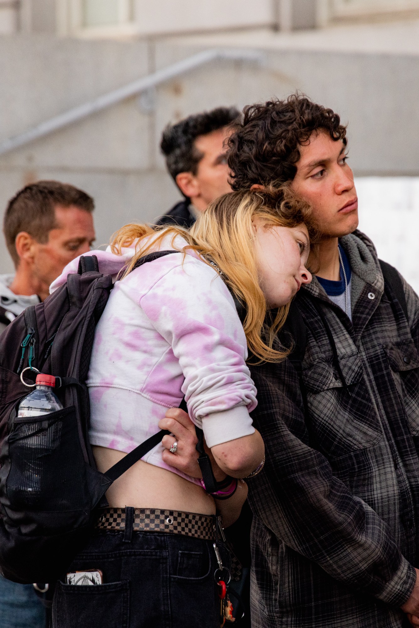 A young woman with a pink tie-dye hoodie rests her head on a man's shoulder. They stand among a group of people, near steps in a city setting.