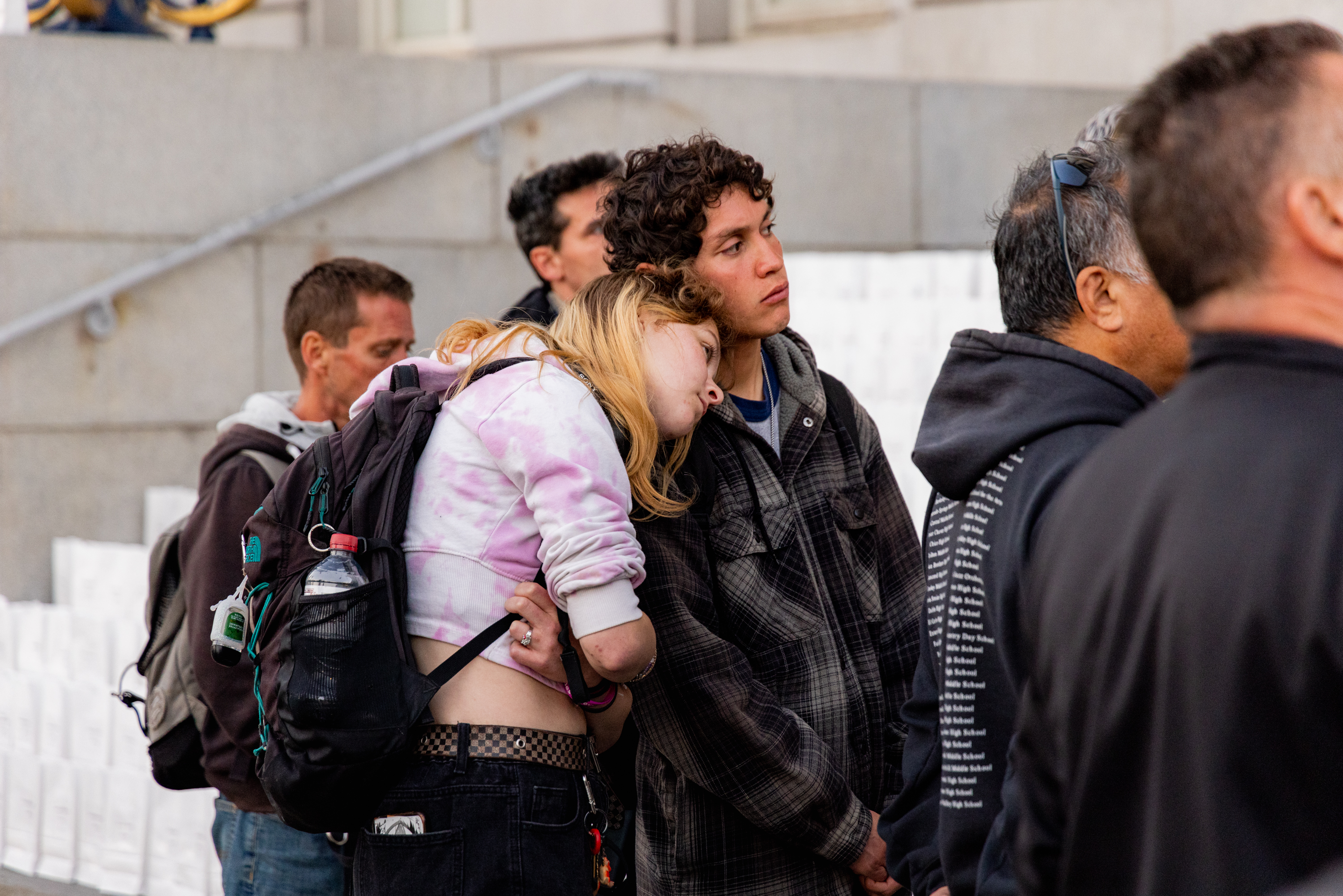 A young woman with a pink tie-dye hoodie rests her head on a man's shoulder. They stand among a group of people, near steps in a city setting.