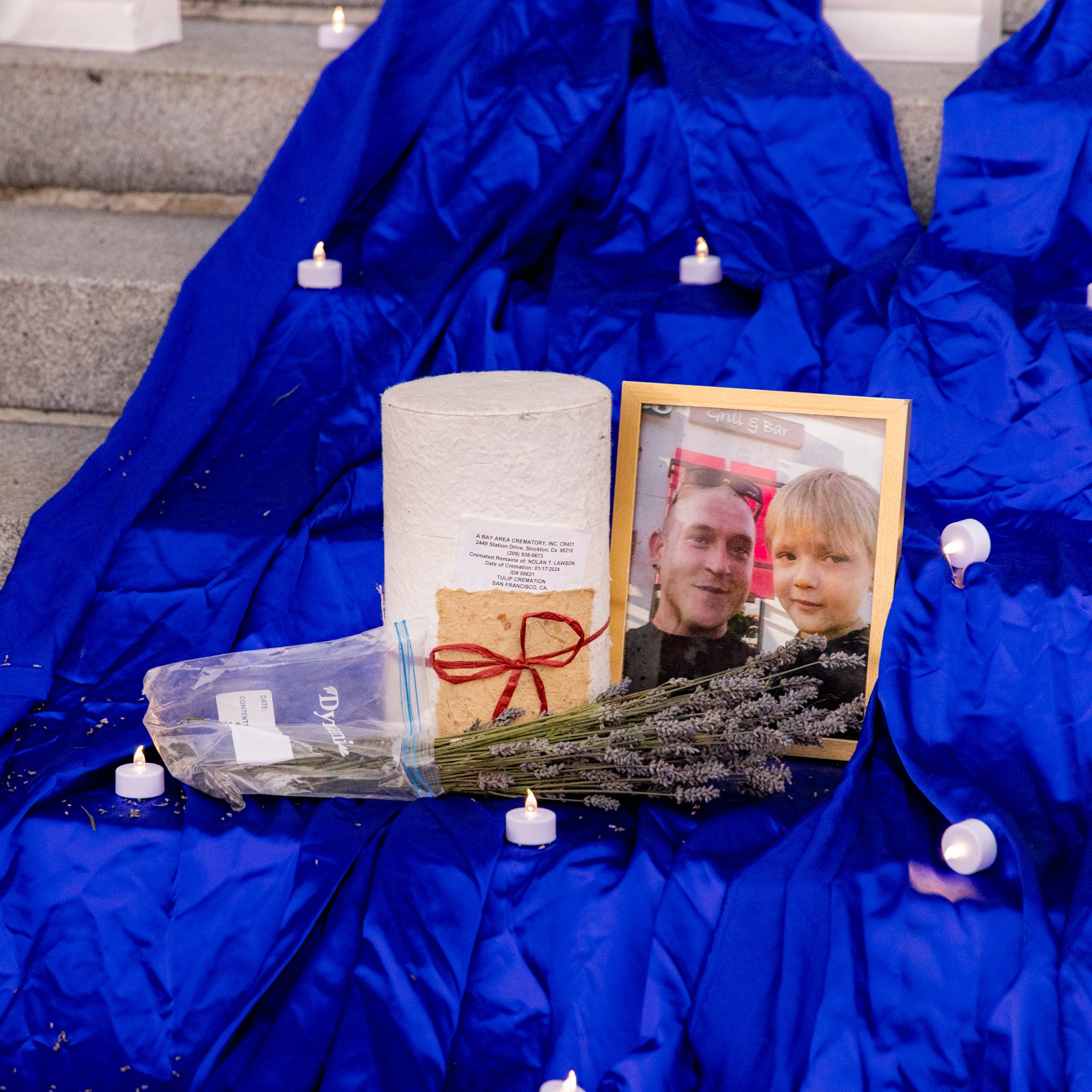 A memorial display on steps features a framed photo of a man and a child, a bouquet of lavender, a cylinder wrapped in white paper, small candles, and a blue cloth.