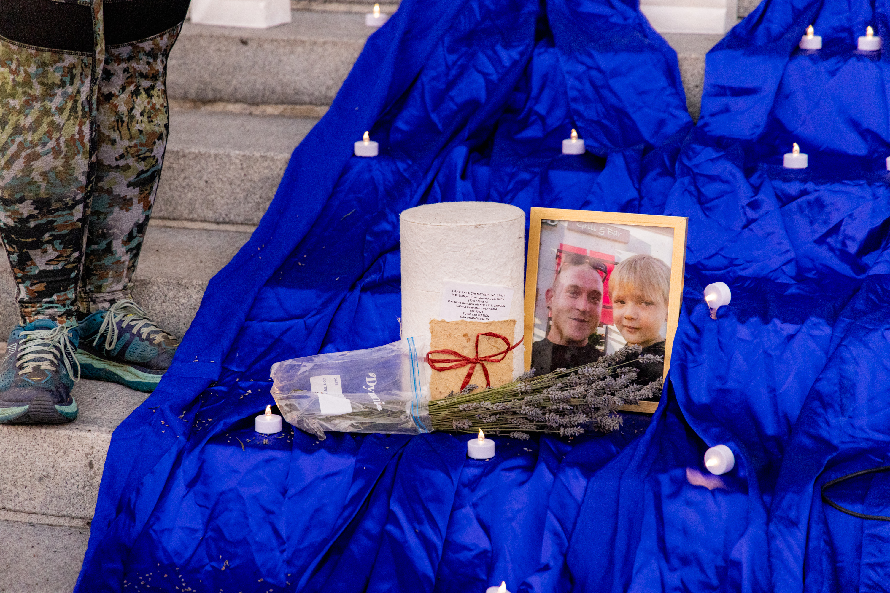 A memorial display on steps features a framed photo of a man and a child, a bouquet of lavender, a cylinder wrapped in white paper, small candles, and a blue cloth.